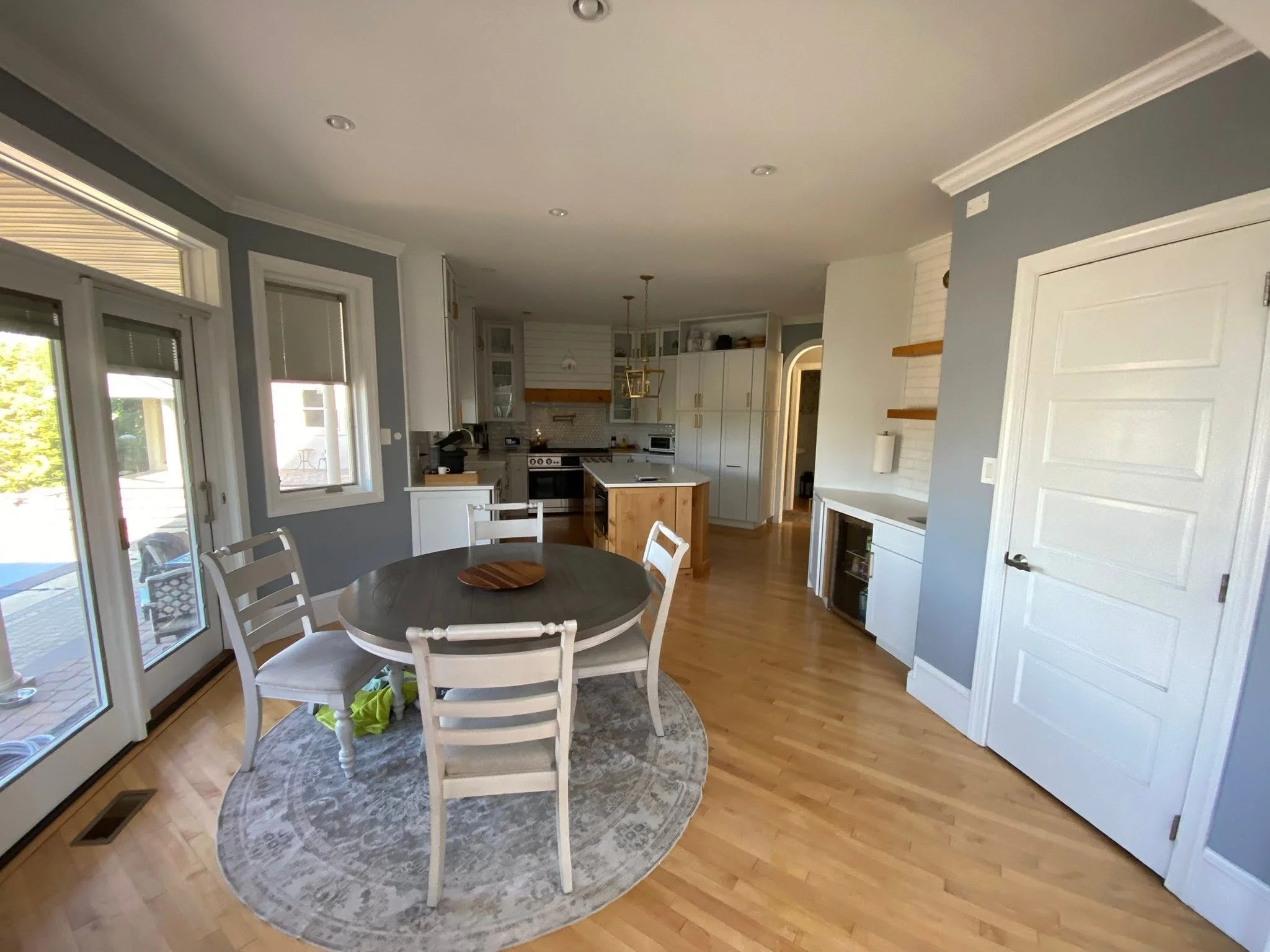 Interior view of a kitchen and dining area with hardwood floors, white cabinets, a small island, and a round dining table with white chairs. Large windows and glass doors let in natural light.