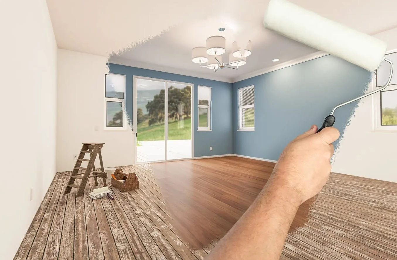 Interior of a room being painted light blue, with a hand using a roller on the wall, and a partially painted wooden floor.