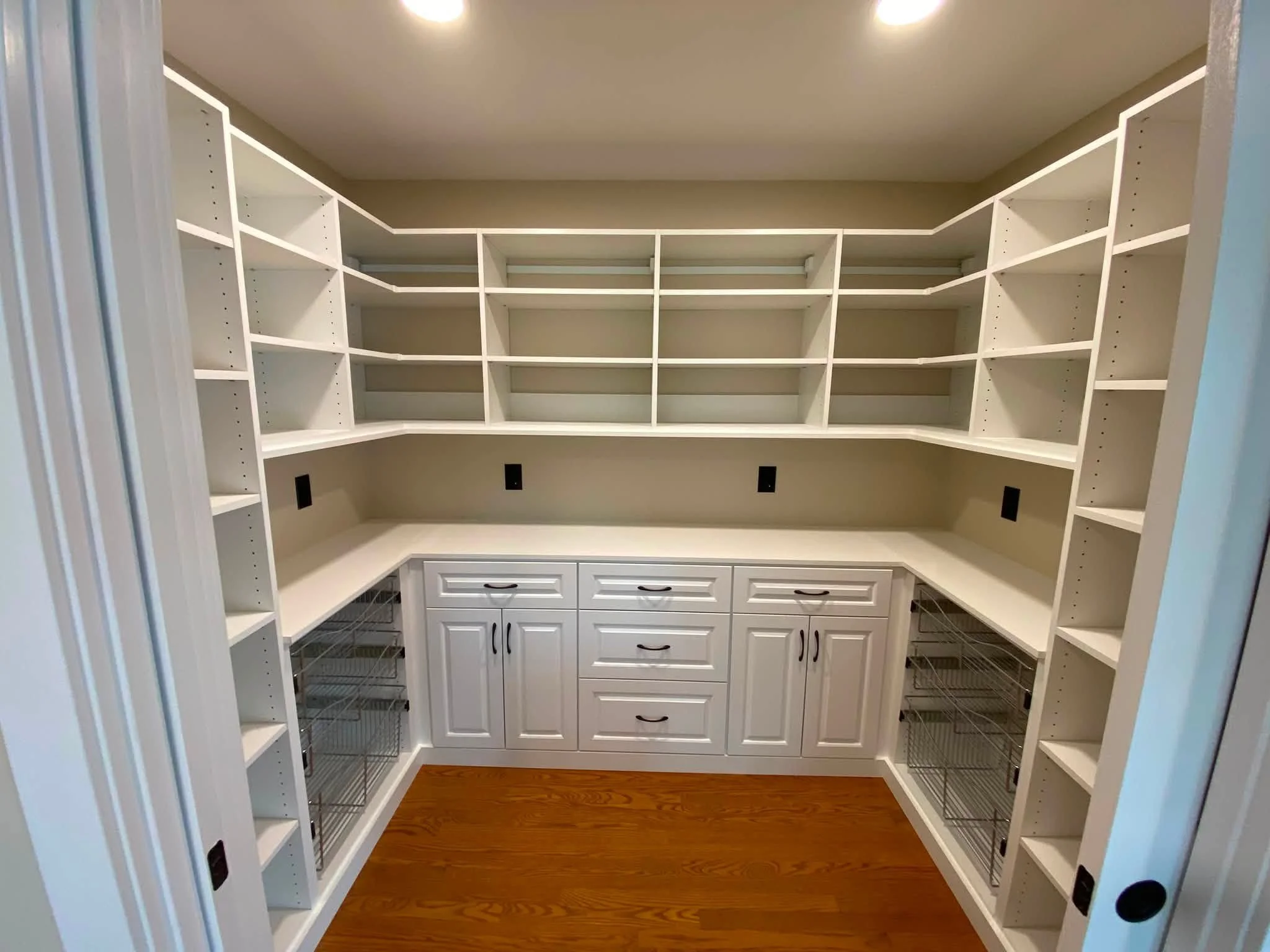 Spacious walk-in closet with white shelving and cabinetry, including drawers and wire baskets, with wood flooring and beige walls.