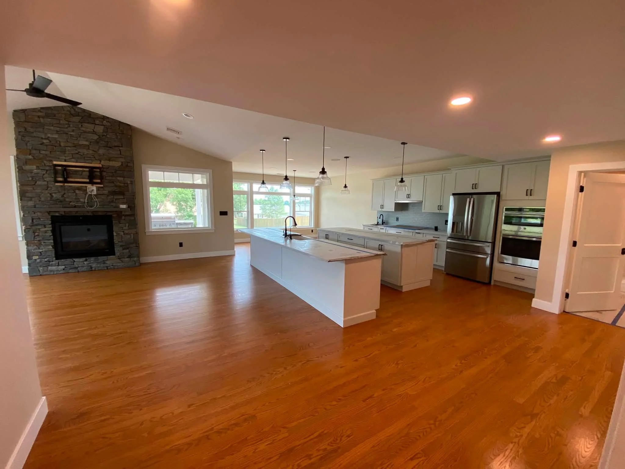 Interior view of a kitchen with wooden floors, white cabinets, a large island with a sink, stainless steel appliances, and a stone fireplace.