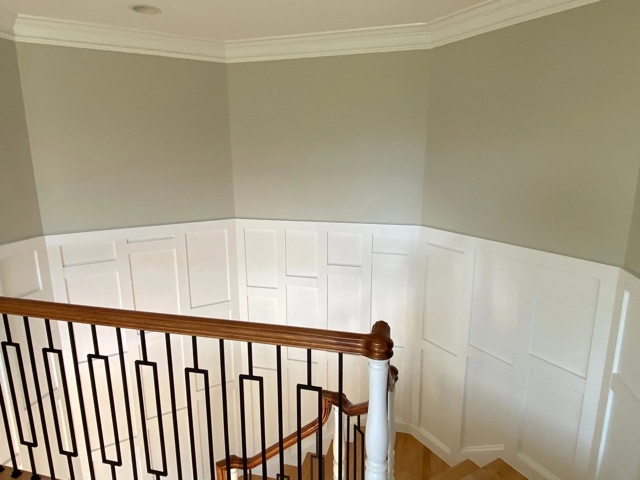 Interior view of a staircase with a wooden handrail and black metal balusters, leading up to a landing with beige cabinets and walls, and a ceiling with a recessed light.
