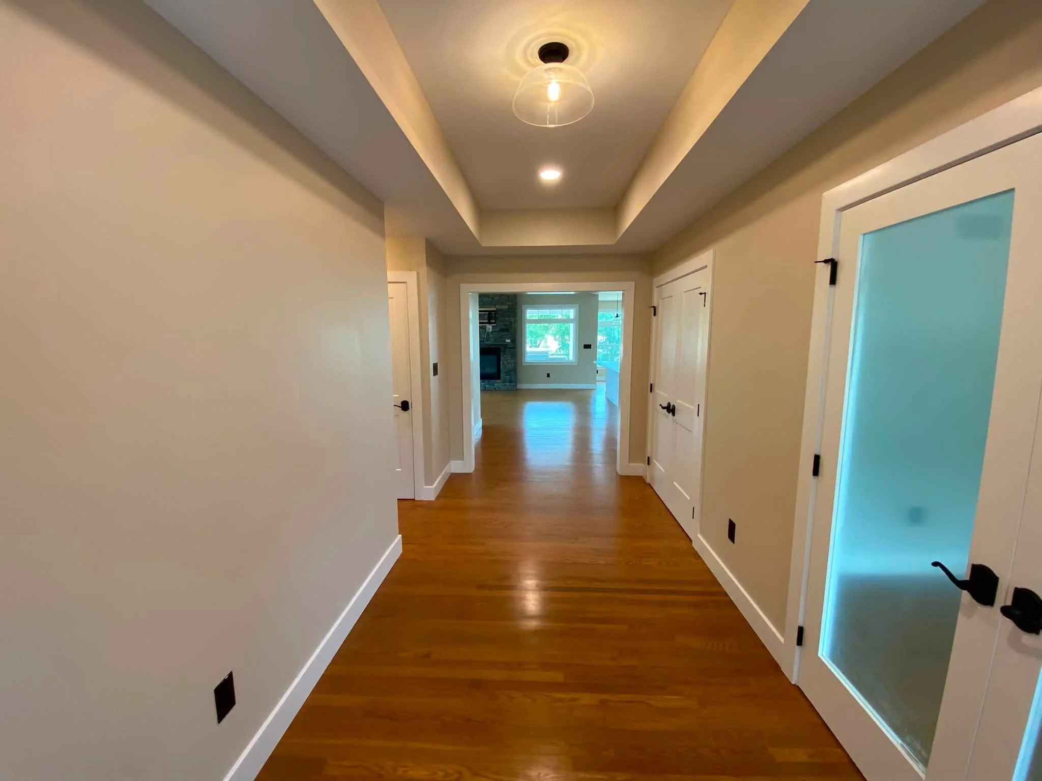 A hallway in a house with blond hardwood flooring, beige walls, white trim, and multiple white doors. The hallway has a recessed ceiling with hidden lighting and a ceiling light fixture. At the end of the hallway, there is a view into a living room a