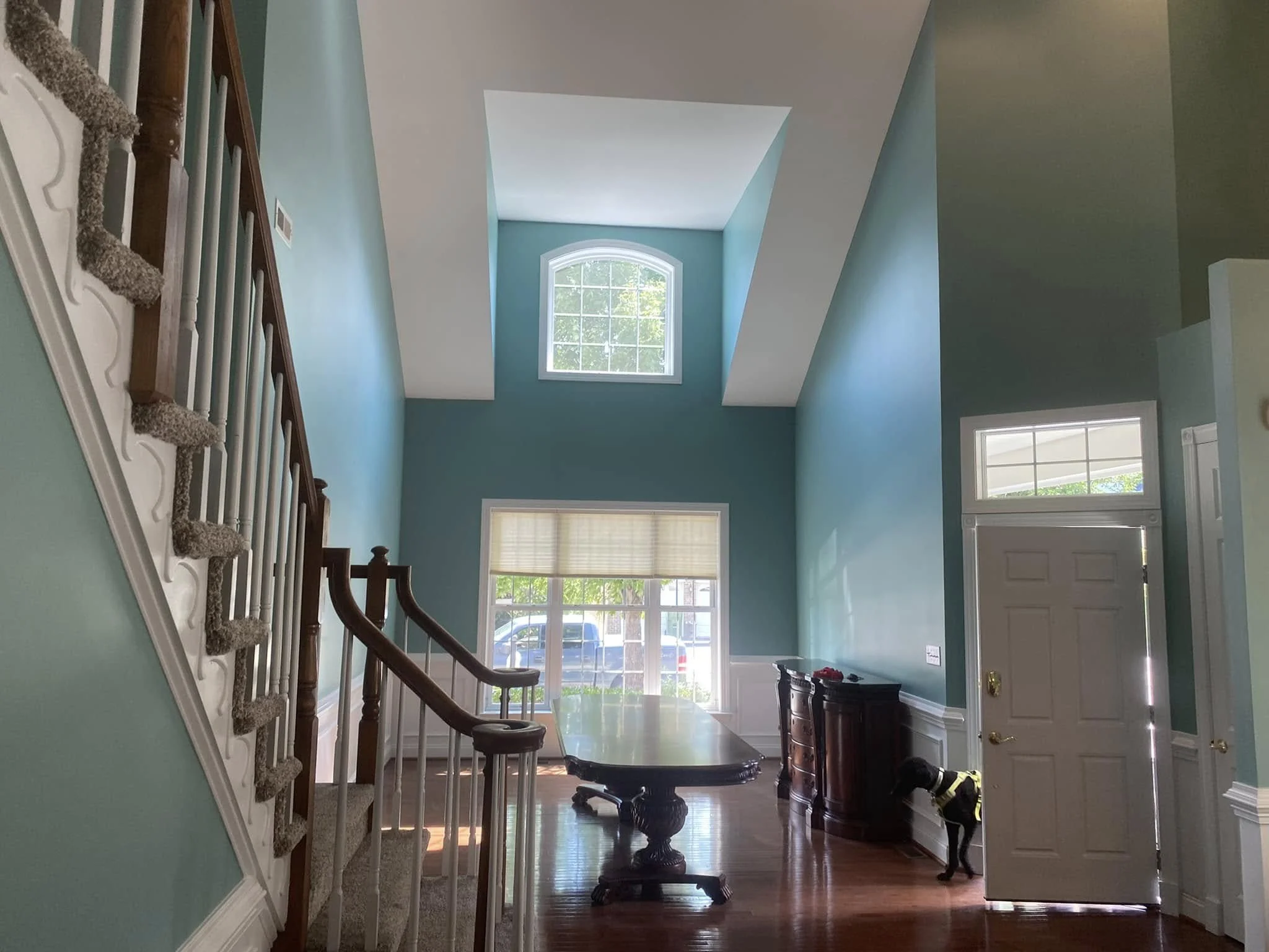 Interior view of a bright hallway with a staircase on the left, a large window with white trim, and a light blue wall color. There is furniture and decorations near the entrance.