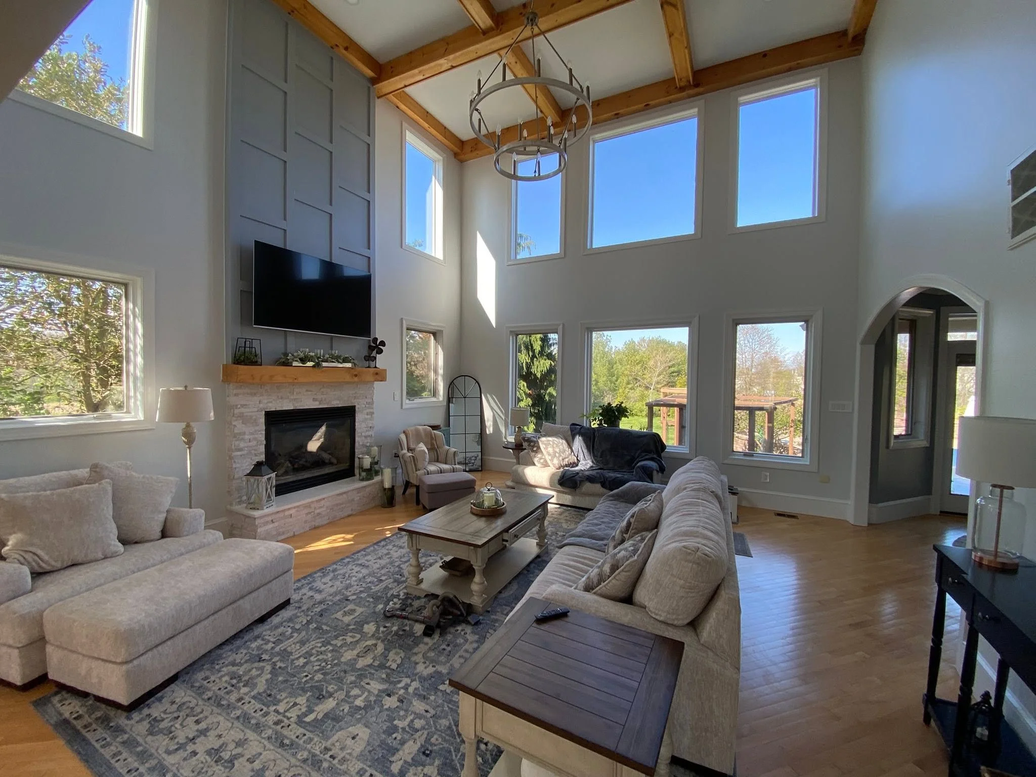 A spacious living room with high ceiling, large windows, and wooden beams. It features a beige sofa, armchair, coffee table, and a fireplace with a wooden mantel decorated with greenery. There is a television mounted above the fireplace and a pattern