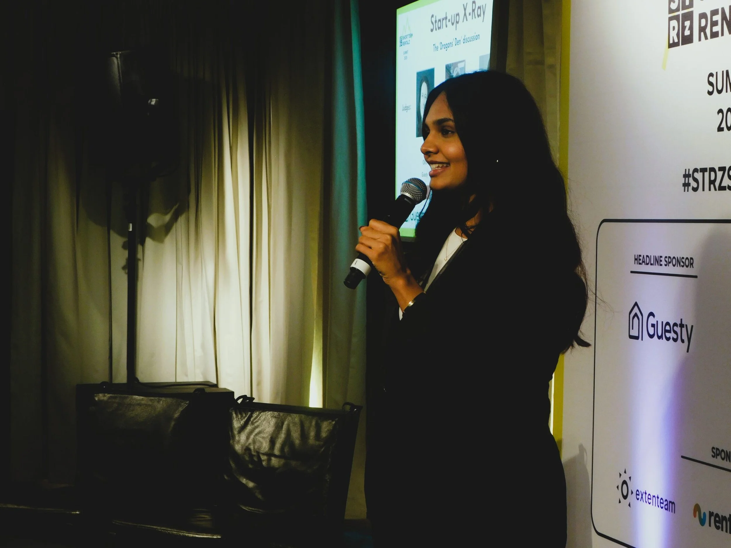 A woman with long dark hair speaking into a microphone at a presentation, with a large screen behind her displaying a slide titled 'Start-up X-Ray' and a discussion on 'The Dragons Den.'