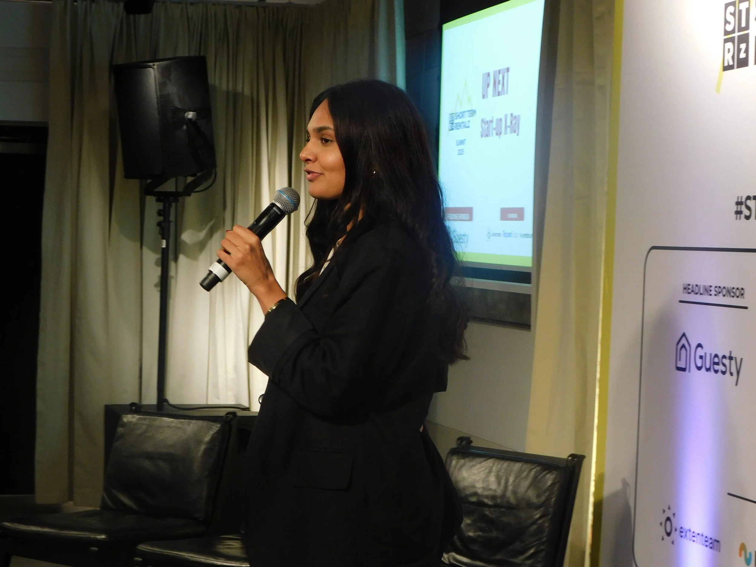 A woman in black speaking into a microphone during a presentation, standing in front of a screen with presentation slides and a banner with logos.