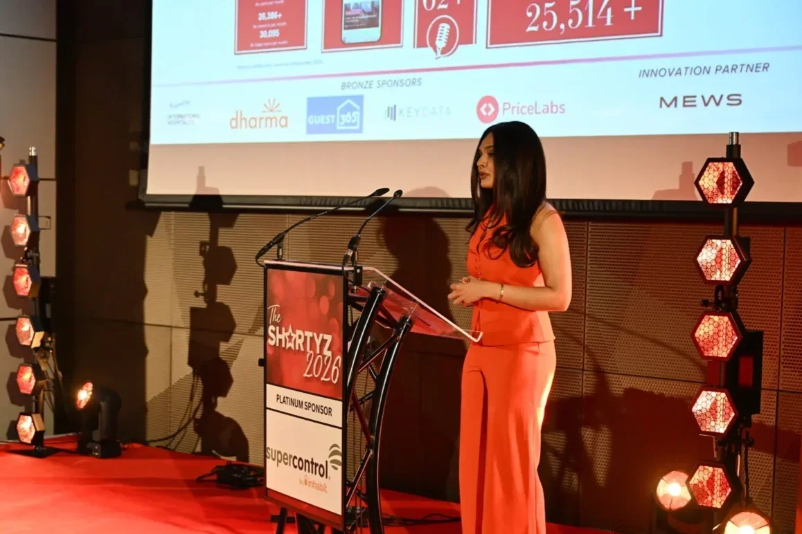 A woman in an orange outfit standing at a podium with a microphone on stage during a conference or event, with a large screen displaying a presentation behind her.