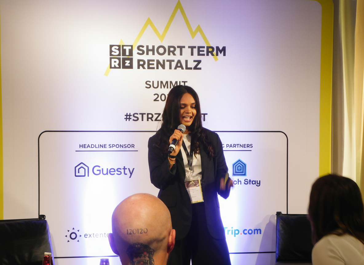 A woman speaking into a microphone at a conference with a large backdrop behind her that reads "Short Term Rentalz Summit".