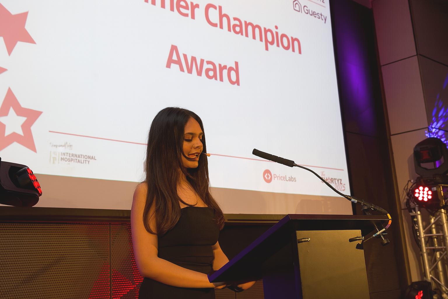 Woman in black dress speaking at a podium during an award ceremony, with a large screen behind her displaying 'Better Champion Award' and event logos.