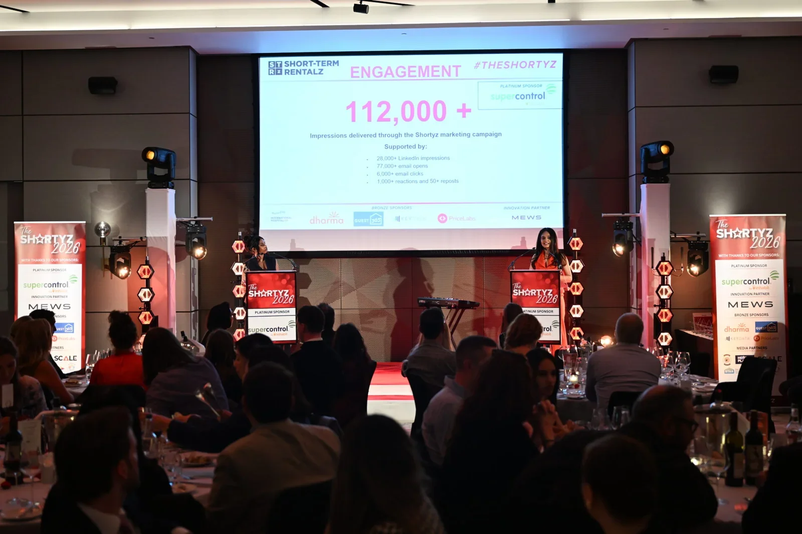 A conference or event with two women speaking at podiums on stage, a large screen behind them displaying presentation data about marketing campaign engagement, audience seated at tables in a dimly lit room, and promotional banners on each side of the stage.