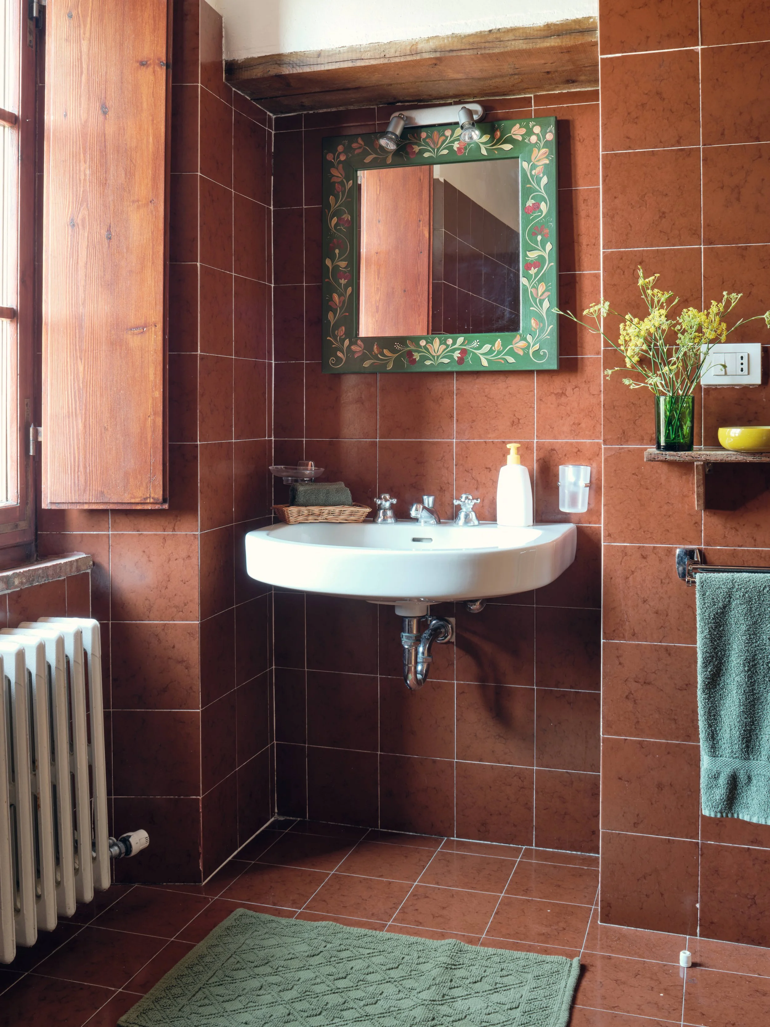 A bathroom with reddish-brown tiled walls and floor, a white oval sink with silver fixtures, a green vase with yellow flowers on a small shelf, a mirror with a decorative green and floral border, a green towel hanging on a rack, a basket with towels,
