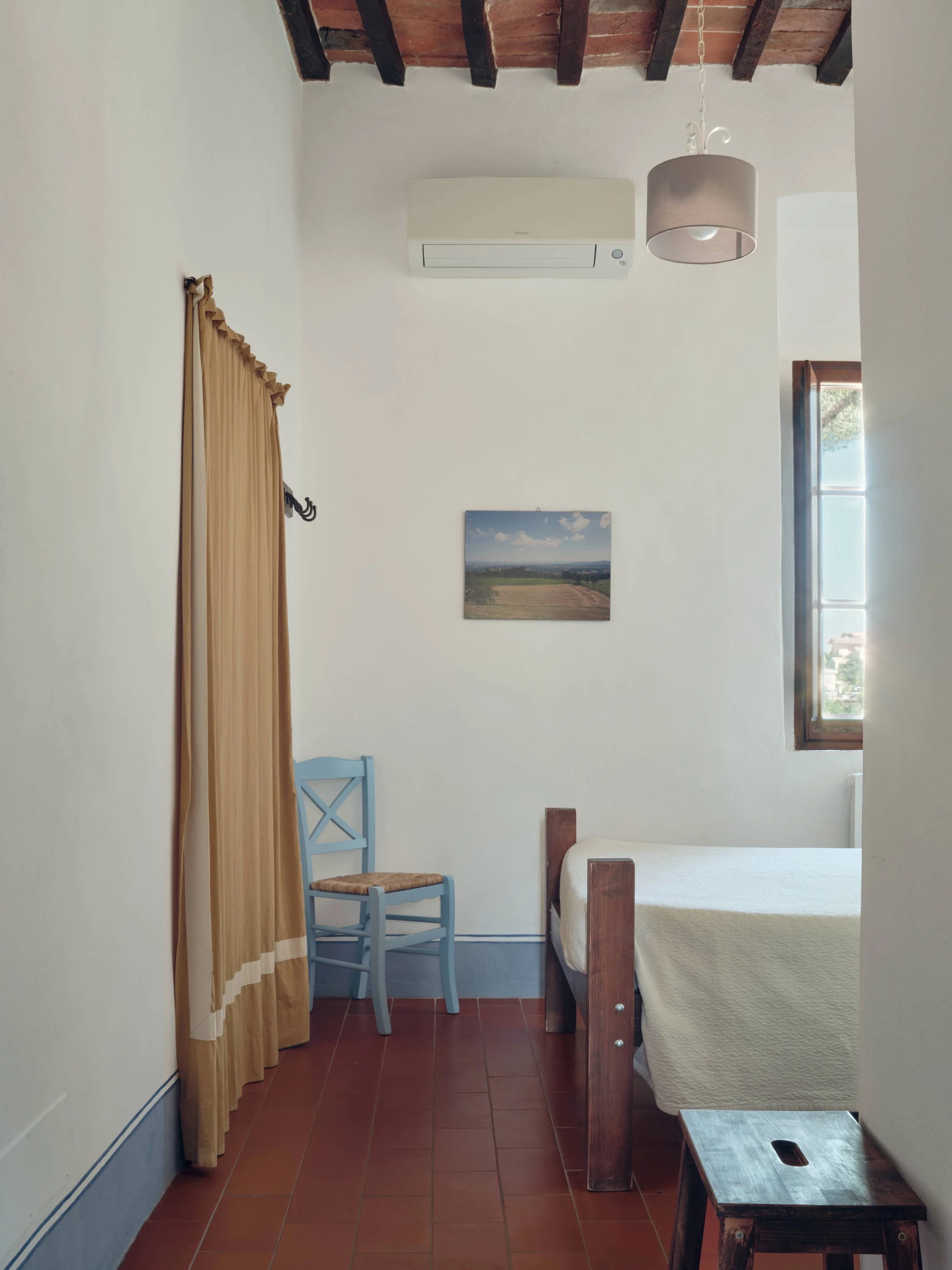 A cozy bedroom with a high ceiling featuring exposed wooden beams and a terracotta tiled floor. There is a single bed with a cream quilt, a small rustic wooden side table at the foot of the bed, a vintage light blue chair with a woven seat, and khaki