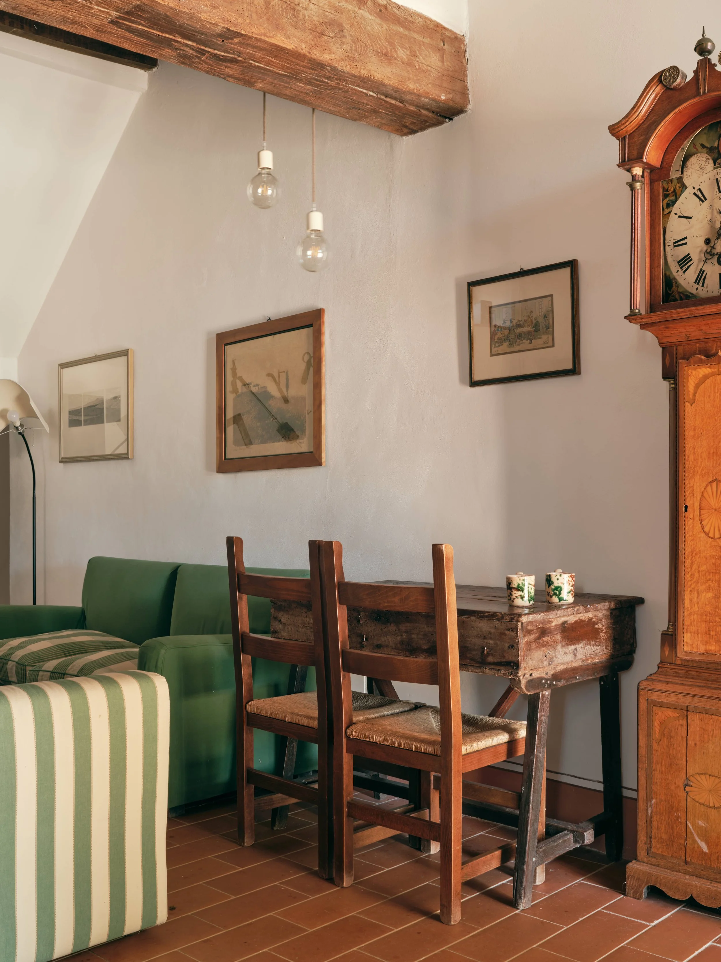 A cozy interior corner featuring a green striped armchair, wooden dining chairs, and a rustic wooden table with two decorative cups. A vintage wooden wall clock, framed pictures, and hanging light bulbs are on the white wall and ceiling with exposed 