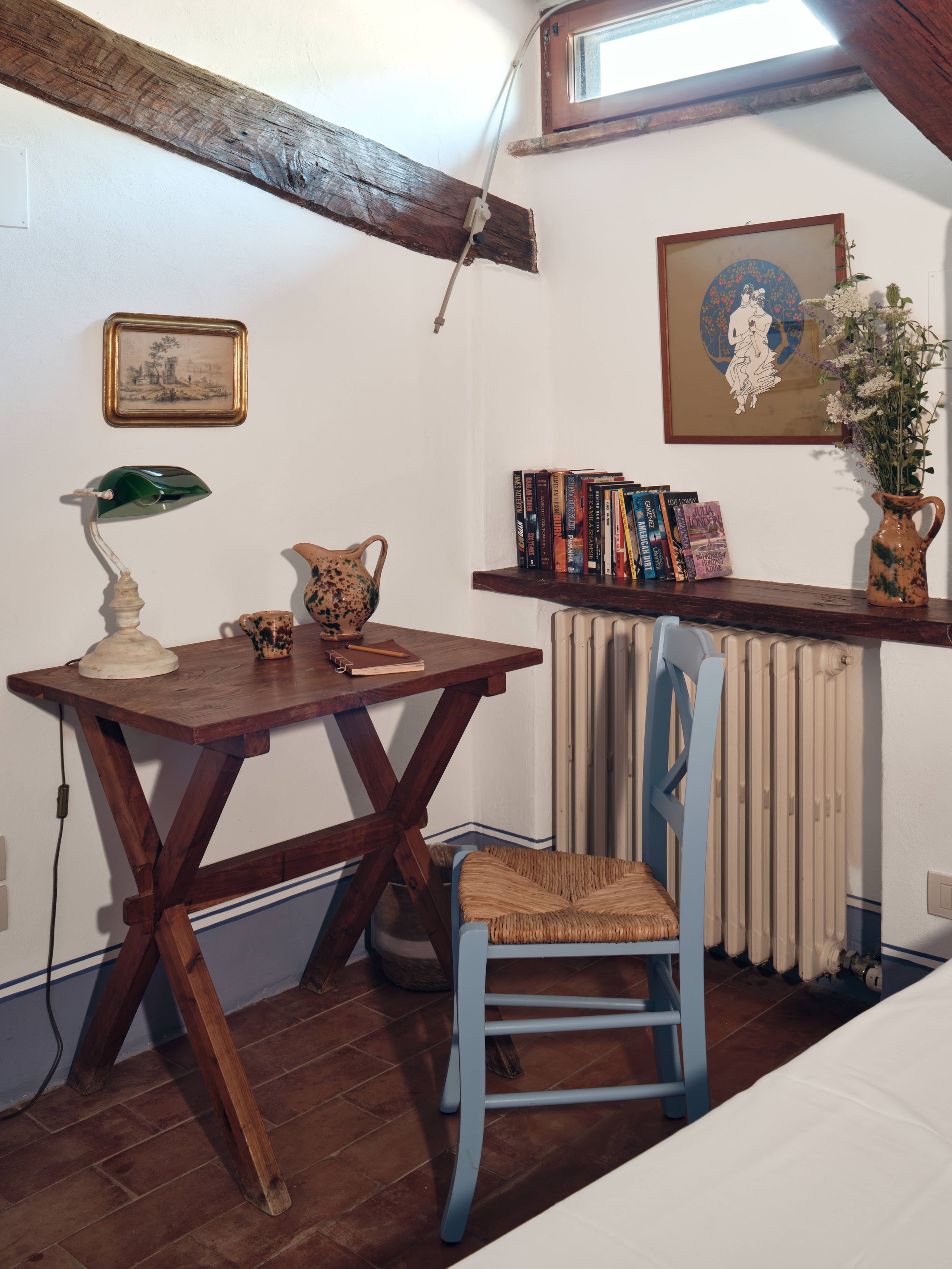 Corner of a room with a wooden desk, blue chair, books, a vase with flowers, wall art, and a window with a wooden beam overhead.