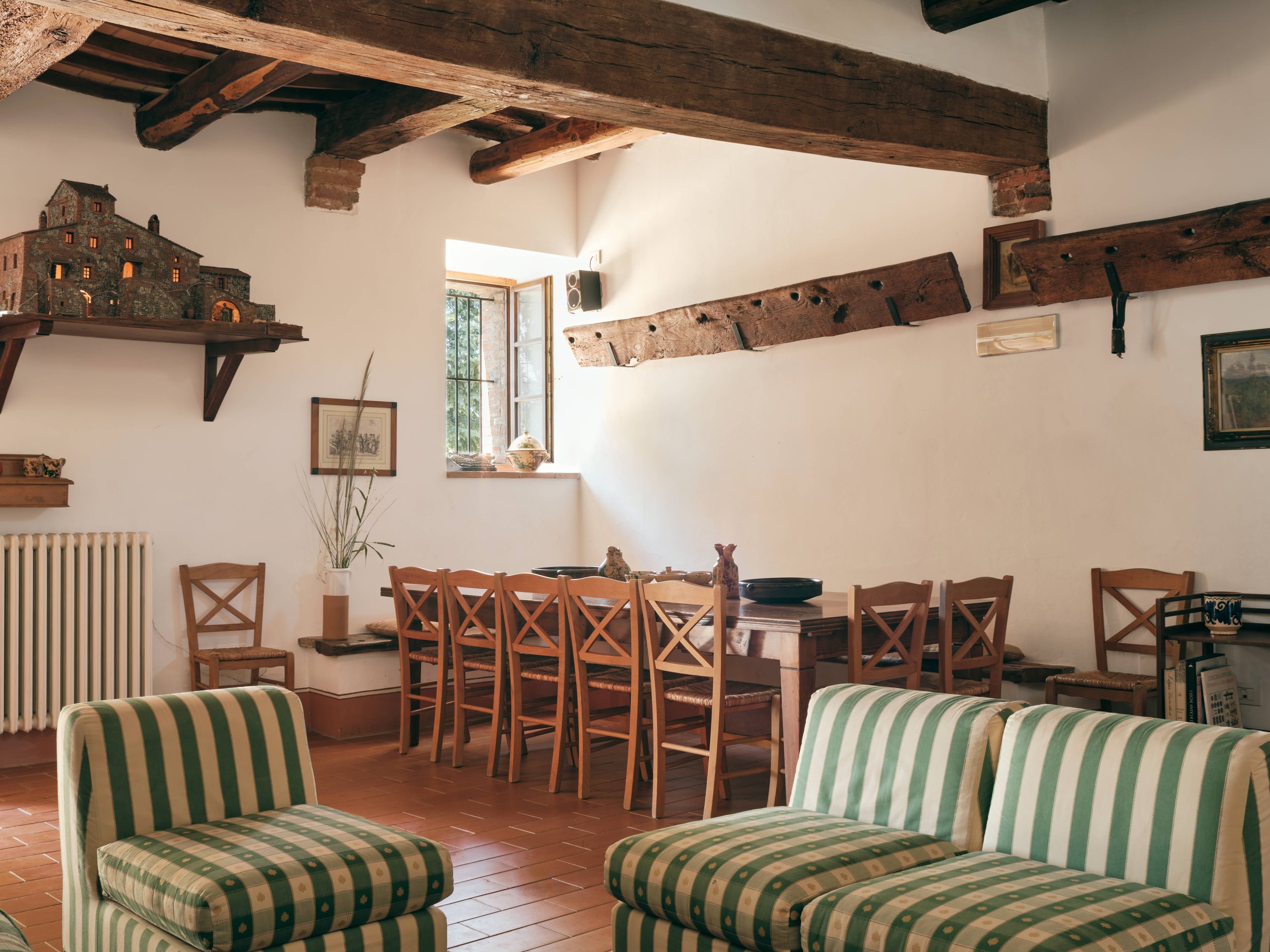 View of a dining room with striped green and white armchairs in the foreground and wooden chairs around a dining table in the background. The room has white walls, exposed wooden beams on the ceiling, and a window letting in natural light. Decor incl