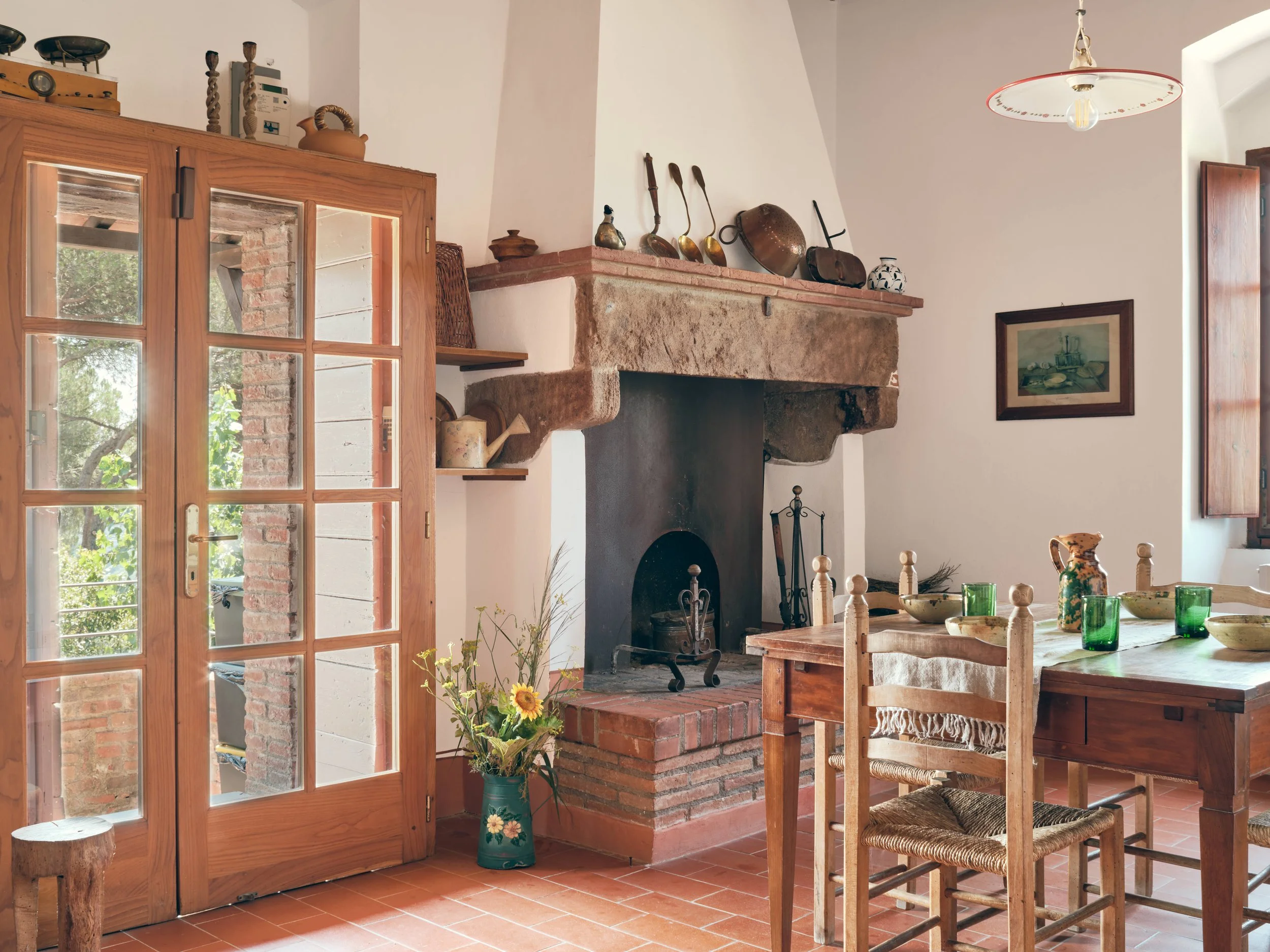 A cozy kitchen with a rustic style, featuring a wooden dining table with woven chairs, a brick-lined fireplace with antique kitchen utensils on the mantel, a glass-fronted door, and a window letting in natural light. Green glassware and a vase with s