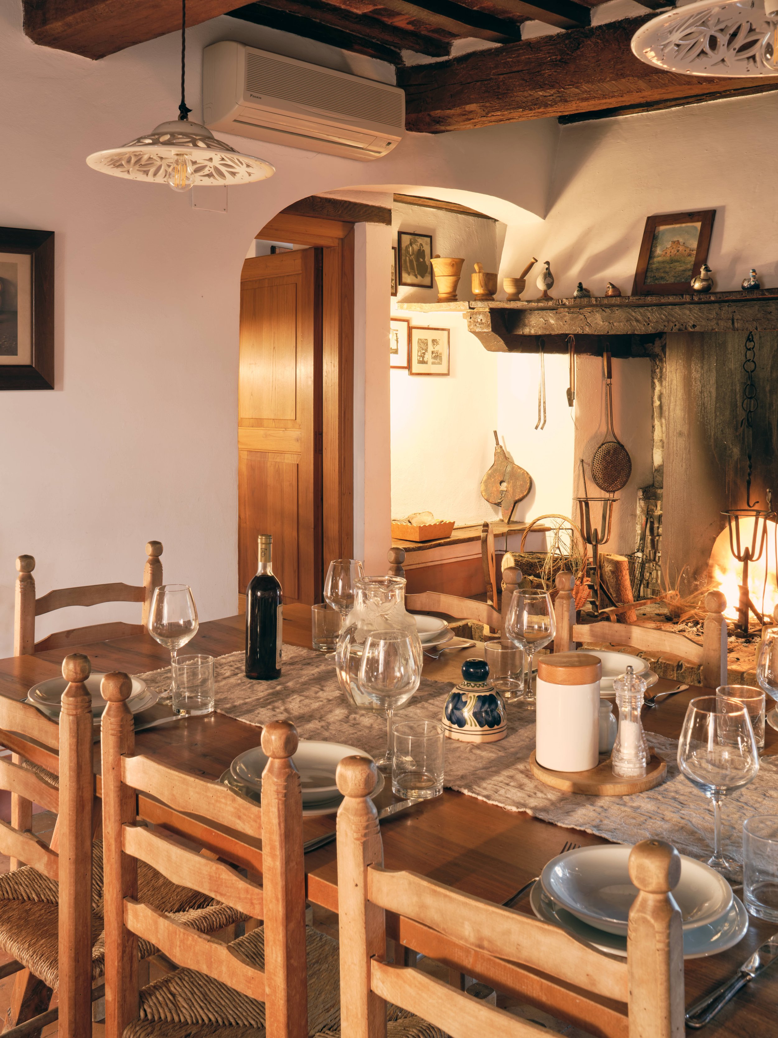 A rustic dining room with a long wooden table set for a meal, including wine bottles, glasses, bowls, and plates. The room features exposed wooden beams, a stone fireplace with various decorative items, and framed artwork on the walls. Hanging pendan