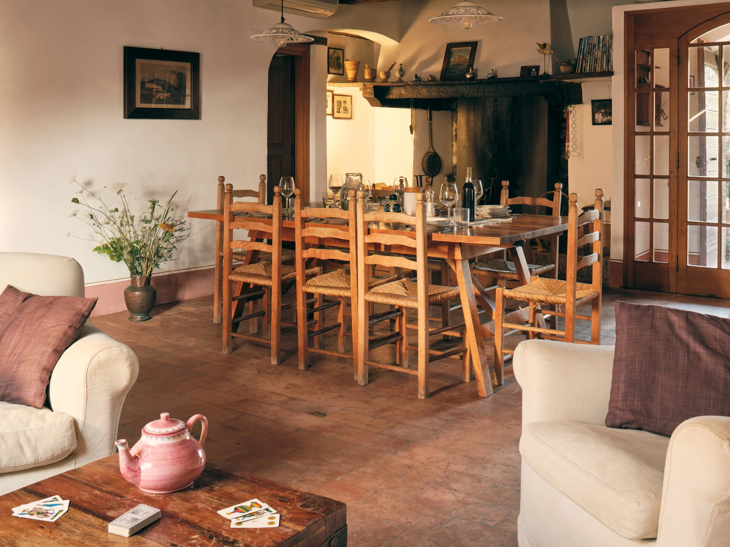 A cozy dining room with a long wooden table set with wine glasses, plates, and a wine bottle. The room has white walls, a stone fireplace with decorative items on the mantle, and wooden chairs around the table. There are large wooden-framed glass doo