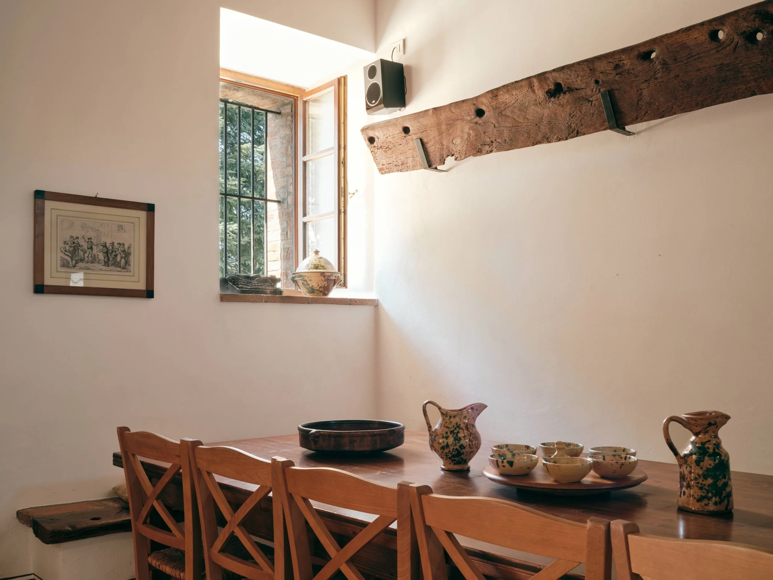 Wooden dining table with ceramic vases and bowls, a window with natural light, a framed artwork on white wall, and a wall-mounted speaker.