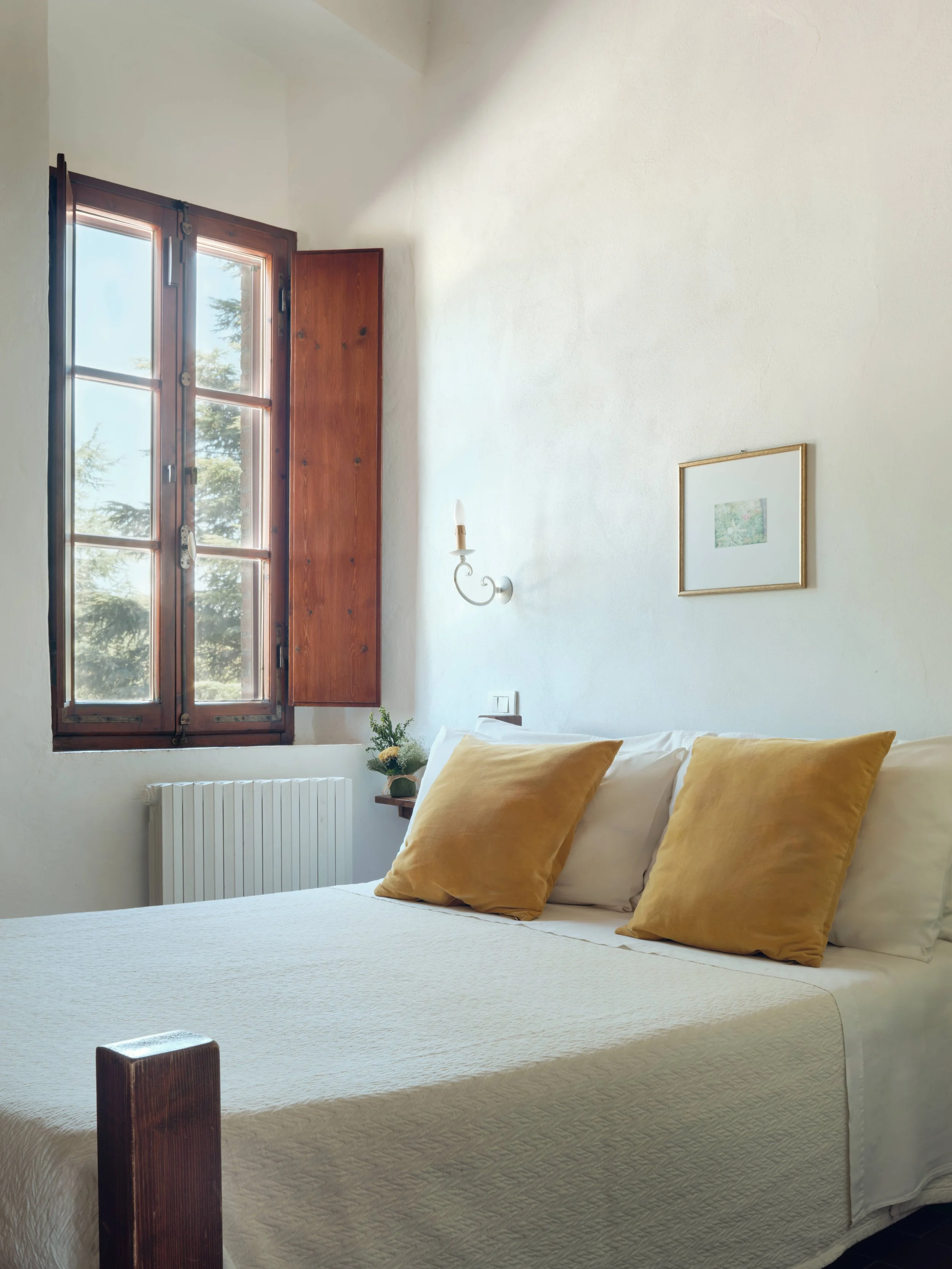 A bedroom with a large window, white wall, framed picture, wall-mounted light fixture, bed with white cover and yellow pillows, and a small side table with a plant.