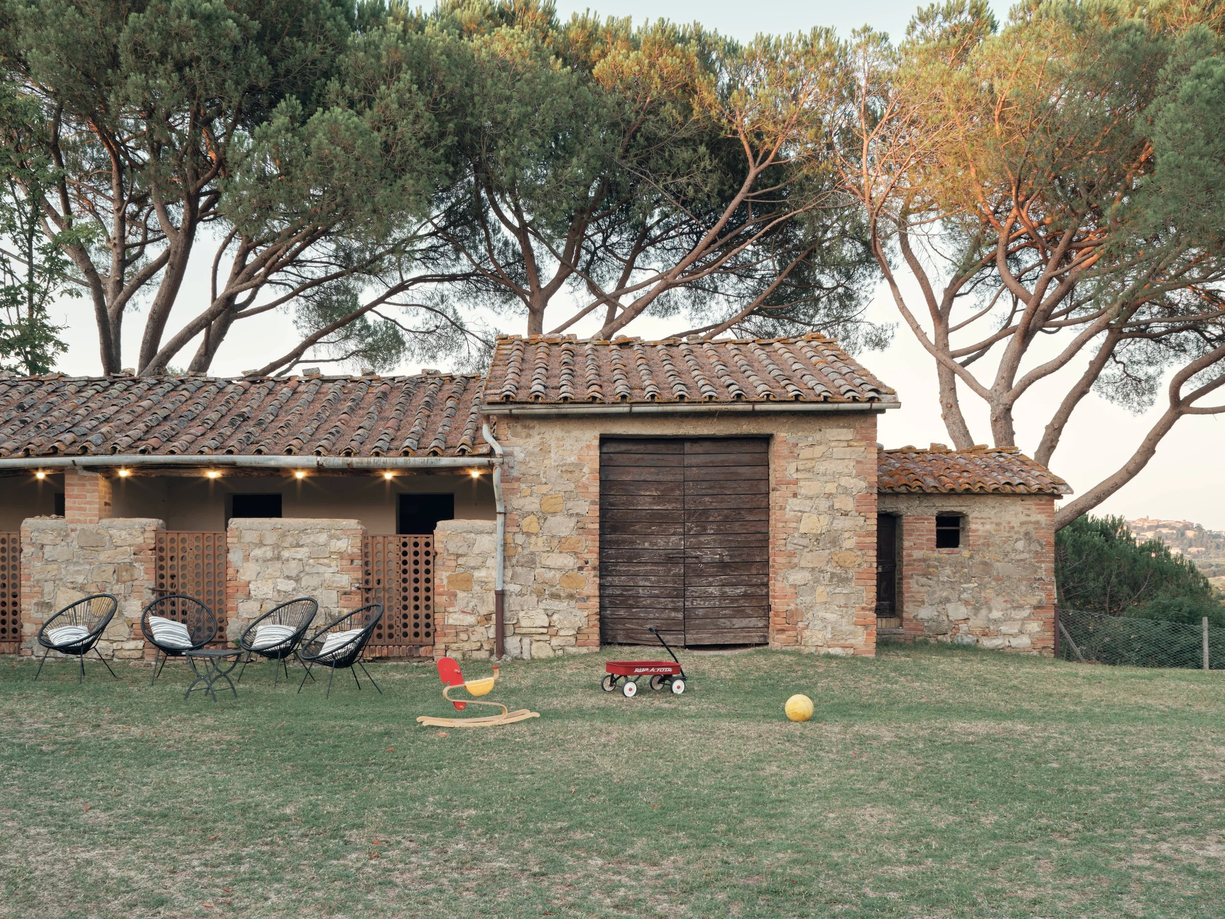 A backyard with a grass lawn, four black chairs with striped cushions, a yellow and red children's rock mixer, a yellow ball, and a red wagon. In the background, there is a stone and brick house with a tiled roof and large trees behind the house.
