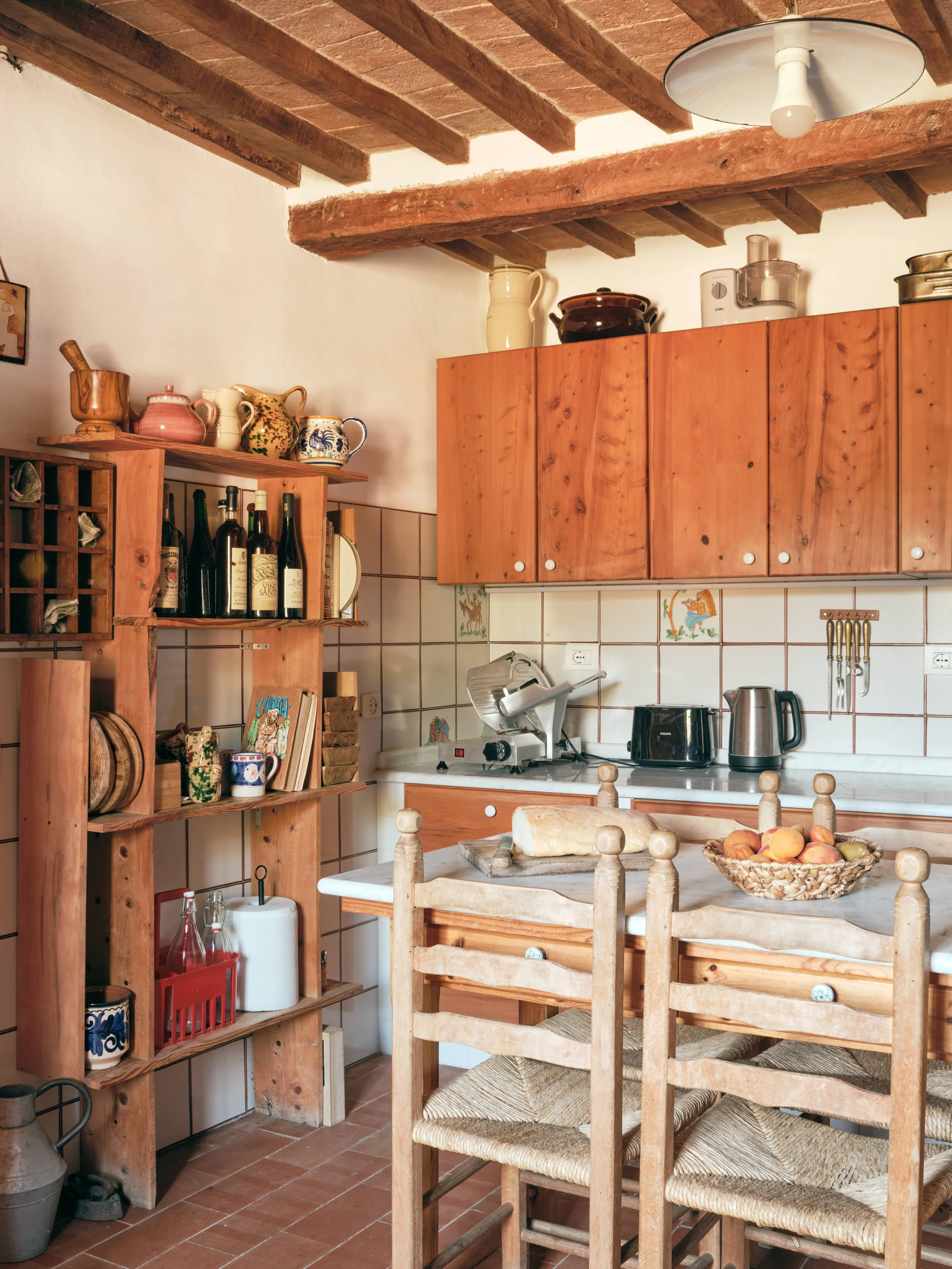 A rustic kitchen with wooden cabinetry, tiled backsplash, and open shelves displaying mugs, bottles, and decorative items. There is a kitchen island with a basket of peaches, a loaf of bread, and wooden chairs around it. The ceiling has exposed woode