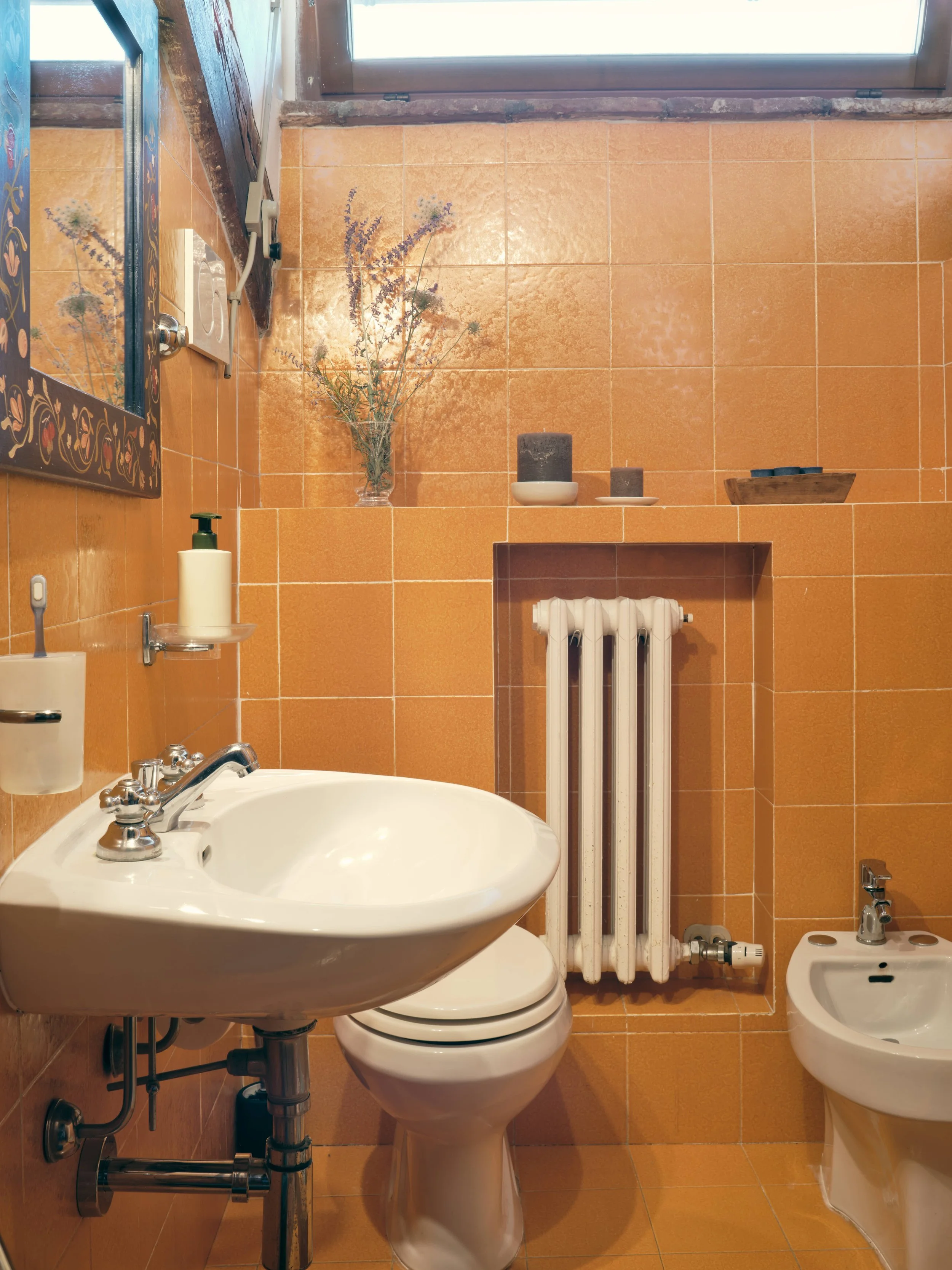 Bathroom with orange tiled walls, a white toilet, a small white sink, a mirror with decorative border, a bidet, and a small shelf with candles, a flower vase, and a window above.