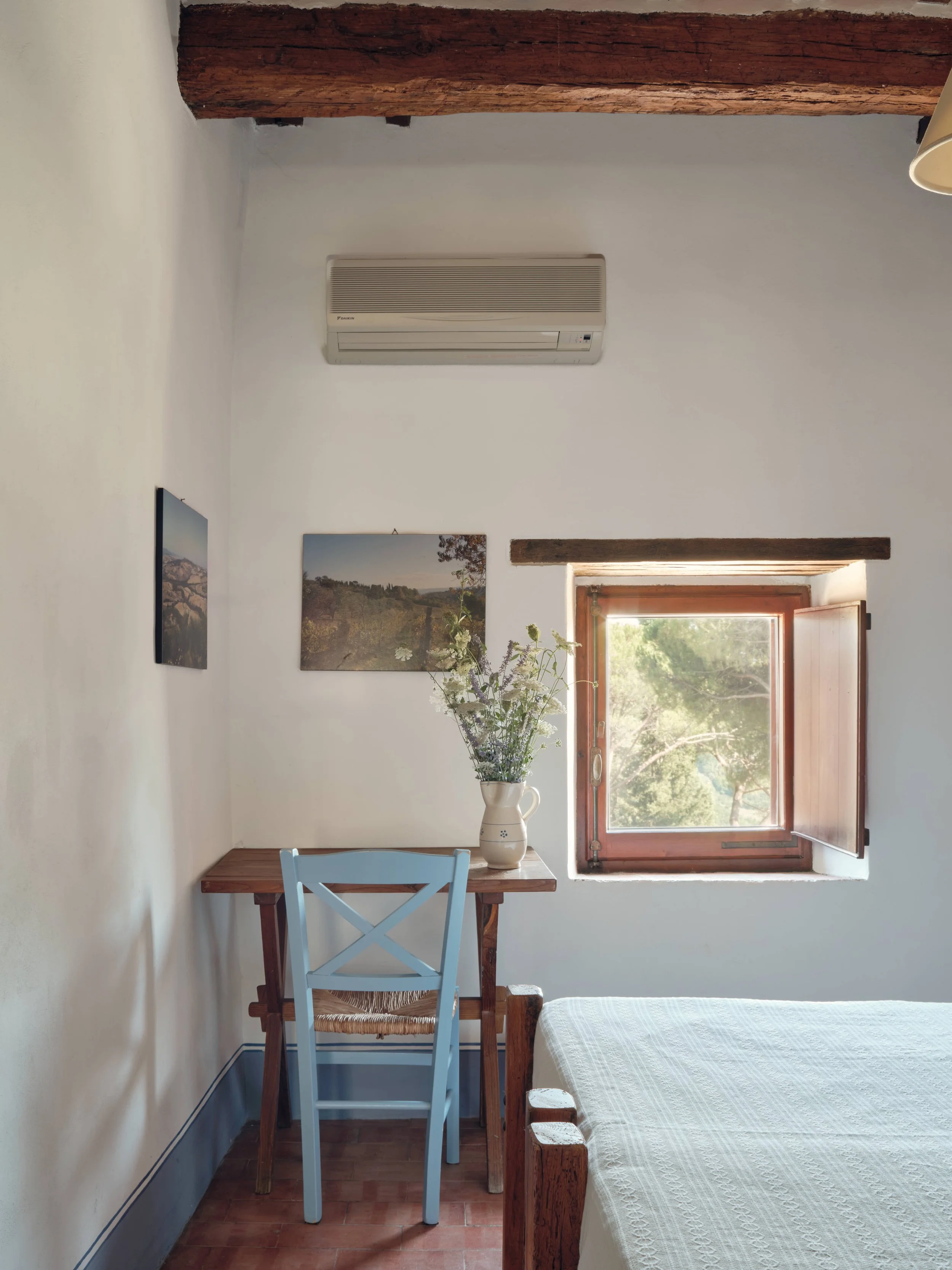 A cozy room with white walls, a window open to a green outdoor view, a wooden table with a blue and a wicker chair, a vase of flowers on the table, and two landscape paintings hung on the wall.
