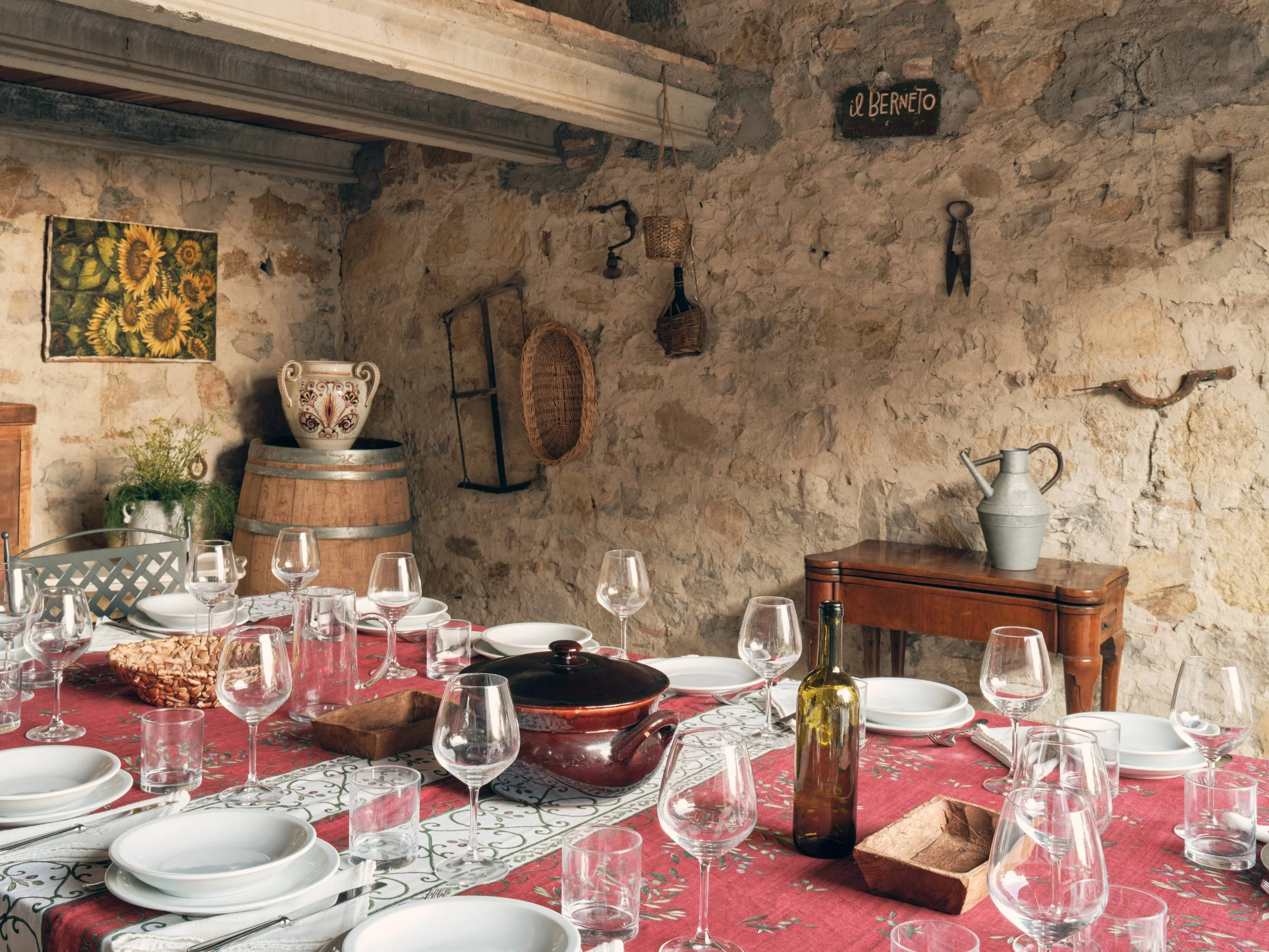 A rustic dining room with a stone wall, decorated with vintage tools, woven baskets, and a sunflower art piece. A table is set with wine glasses, white bowls, a cutting board, a bottle of olive oil, and a covered dish, all on a red and white tableclo