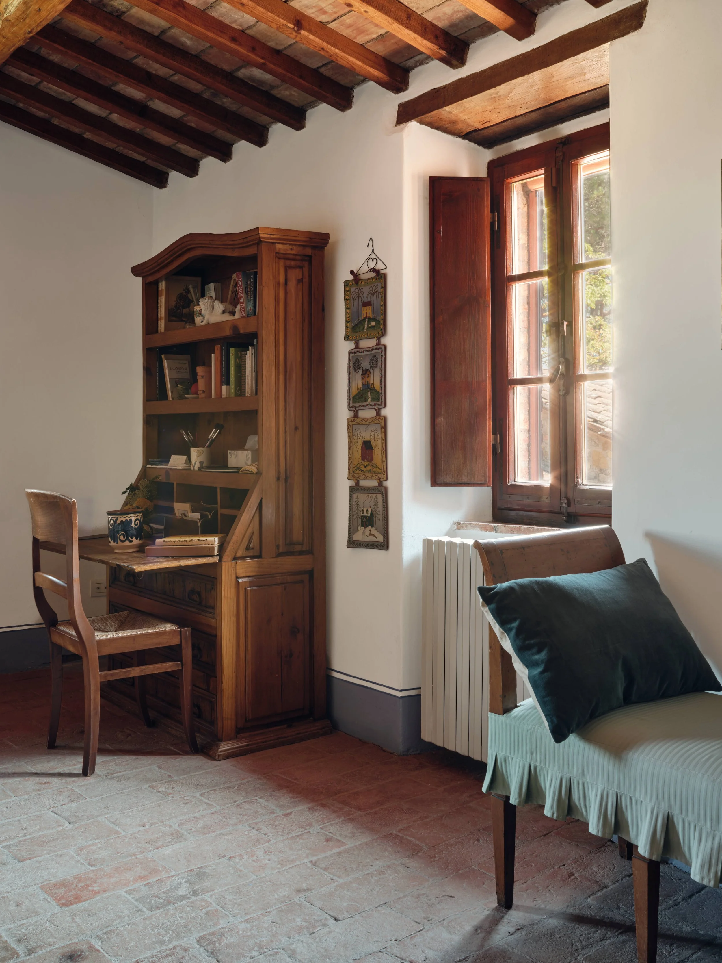 A cozy room with rustic decor featuring a wooden bookshelf filled with books and decorations, a wooden chair, a window with open wooden shutters allowing sunlight in, and a bed with a dark-colored pillow and a green bed skirt, all with a terracotta t