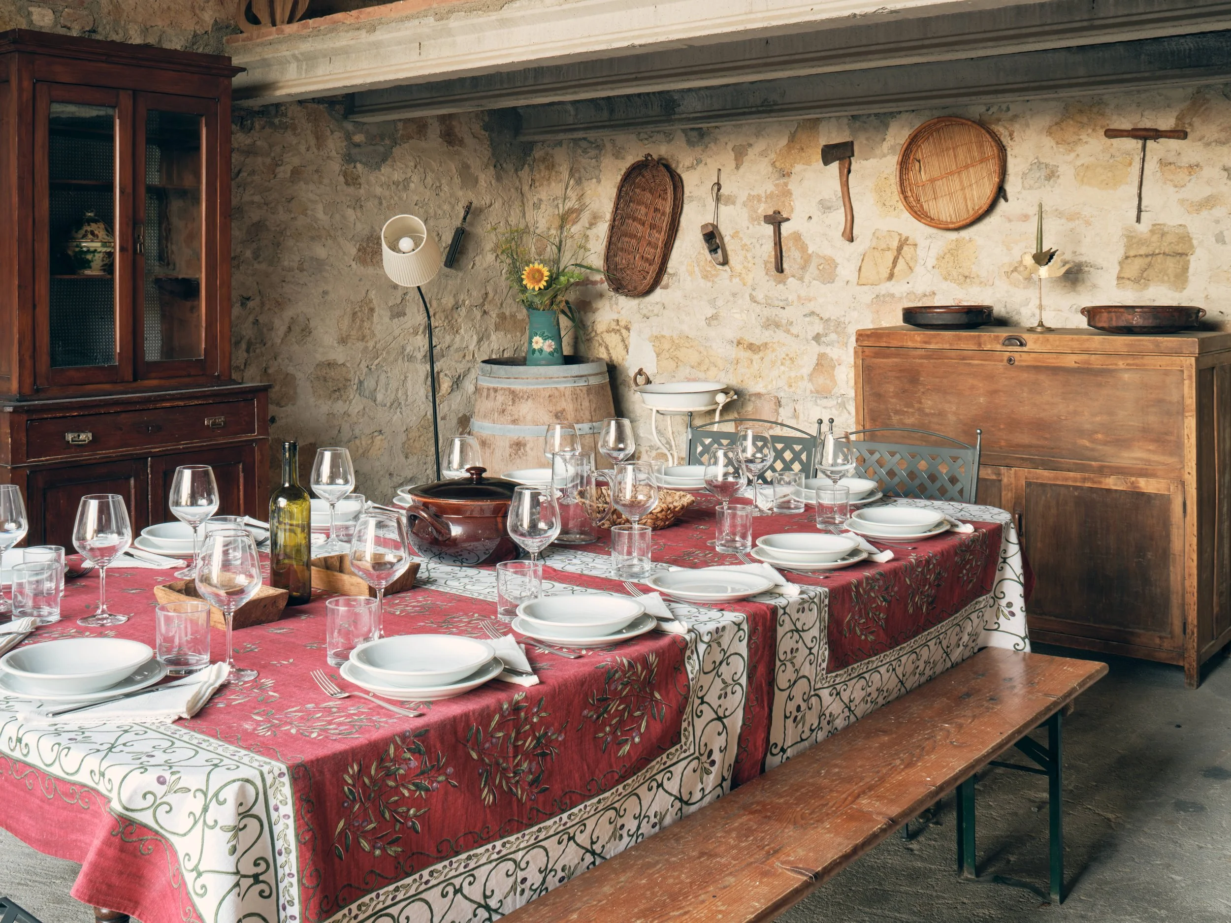 A rustic dining room scene with a long table covered in a red and white patterned tablecloth. The table is set with white dinner plates, wine glasses, and a few bottles of wine. The background features a stone wall with various vintage tools and wove