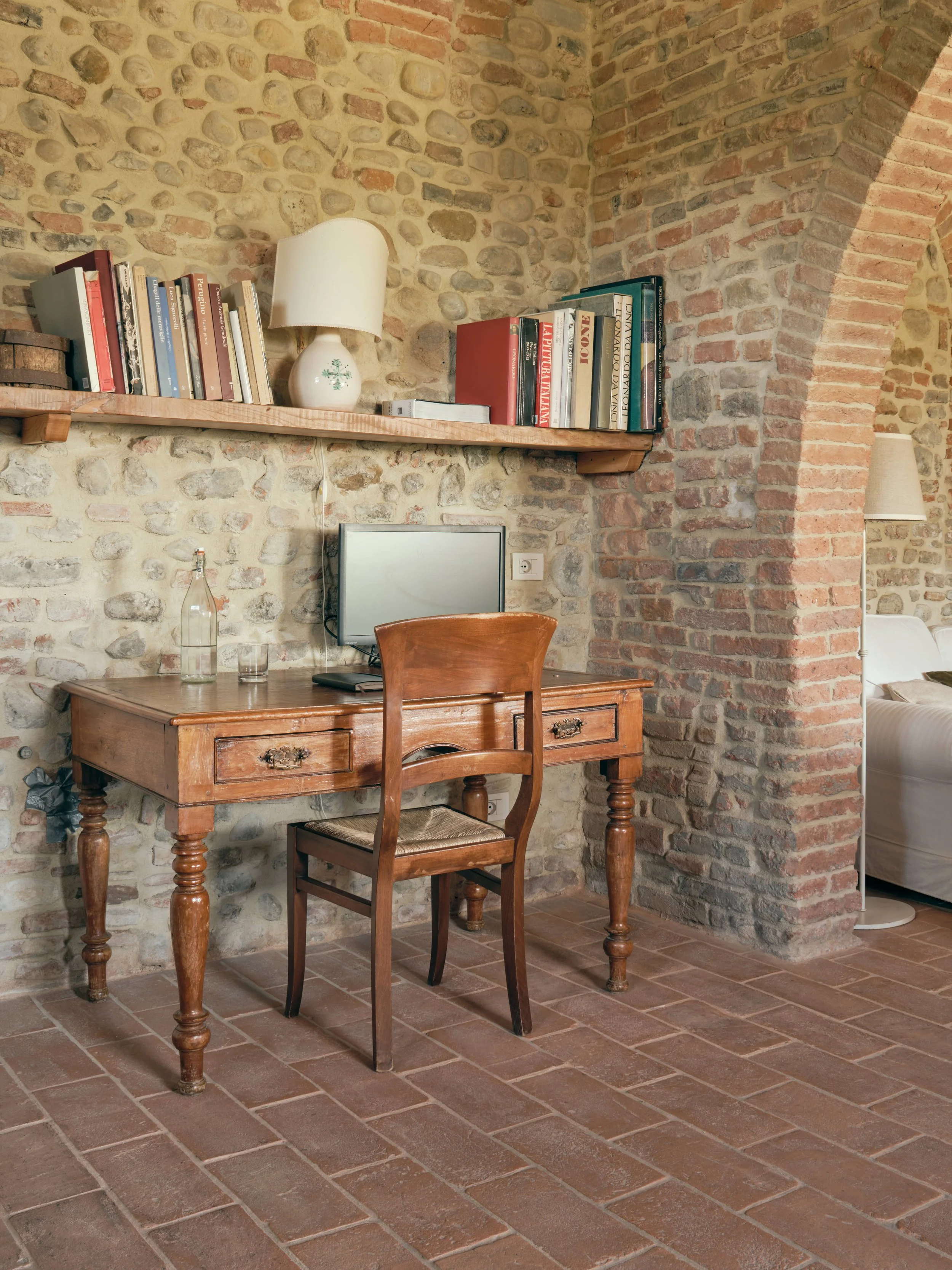 A rustic workspace with a wooden desk and chair set against a brick and stone wall. The desk has a small computer monitor, glass bottle, and glass cup, with a floating shelf above holding books and a table lamp.