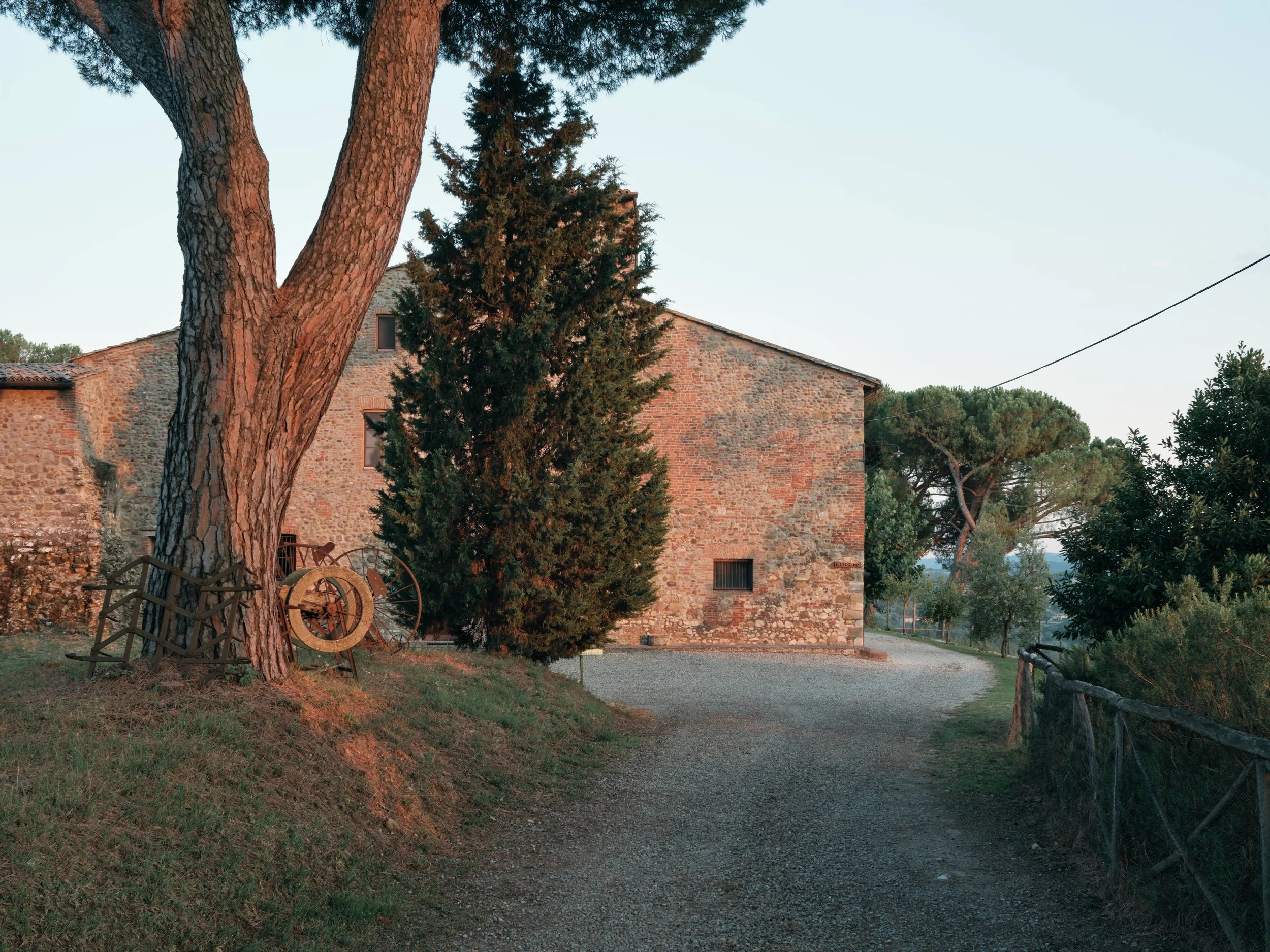 A rustic brick building with small windows, surrounded by tall pine trees, with a dirt and gravel pathway leading towards the building and a cloudy sky overhead.
