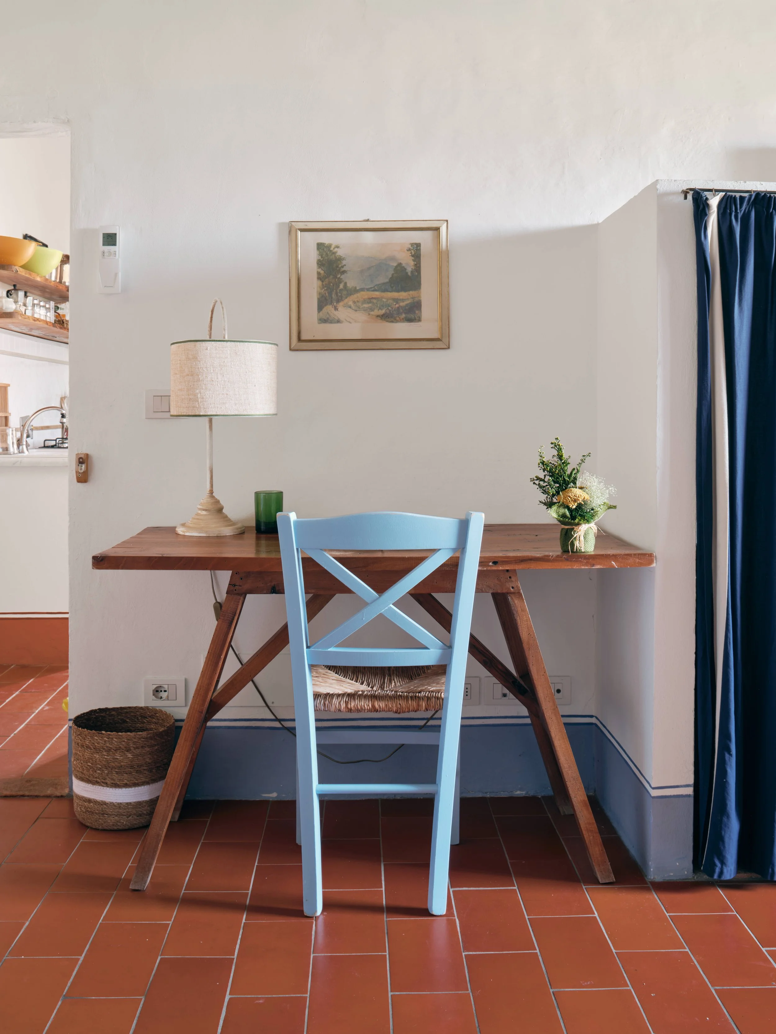 A wooden desk with a blue chair in front, a cream-colored table lamp to the left, and a green candle holder. A bouquet of flowers in a small basket is on the right corner of the desk. Behind the desk, the wall has a framed landscape painting, and the