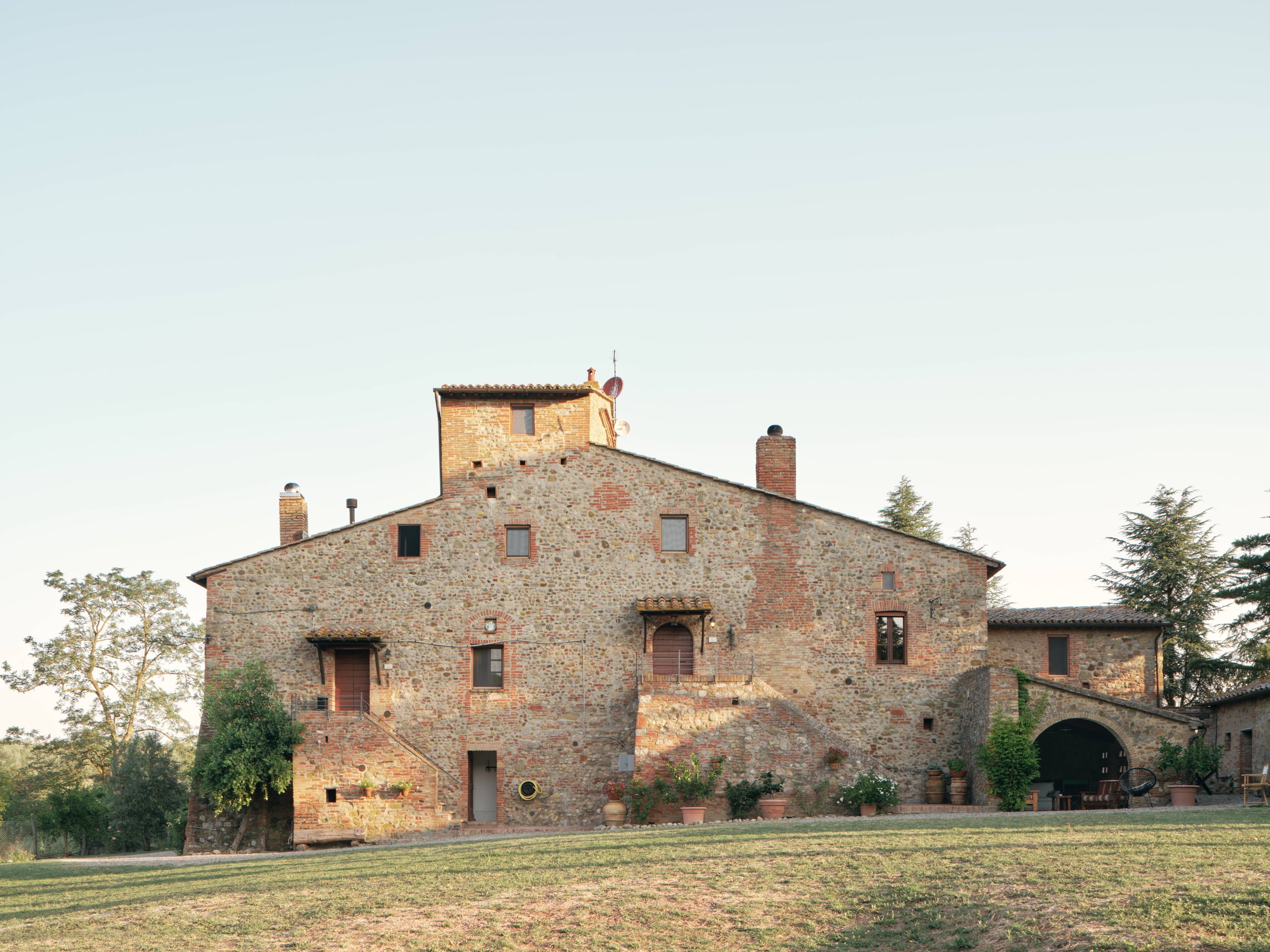 A historic stone and brick building with small windows, surrounded by trees and a grassy yard under a clear sky.