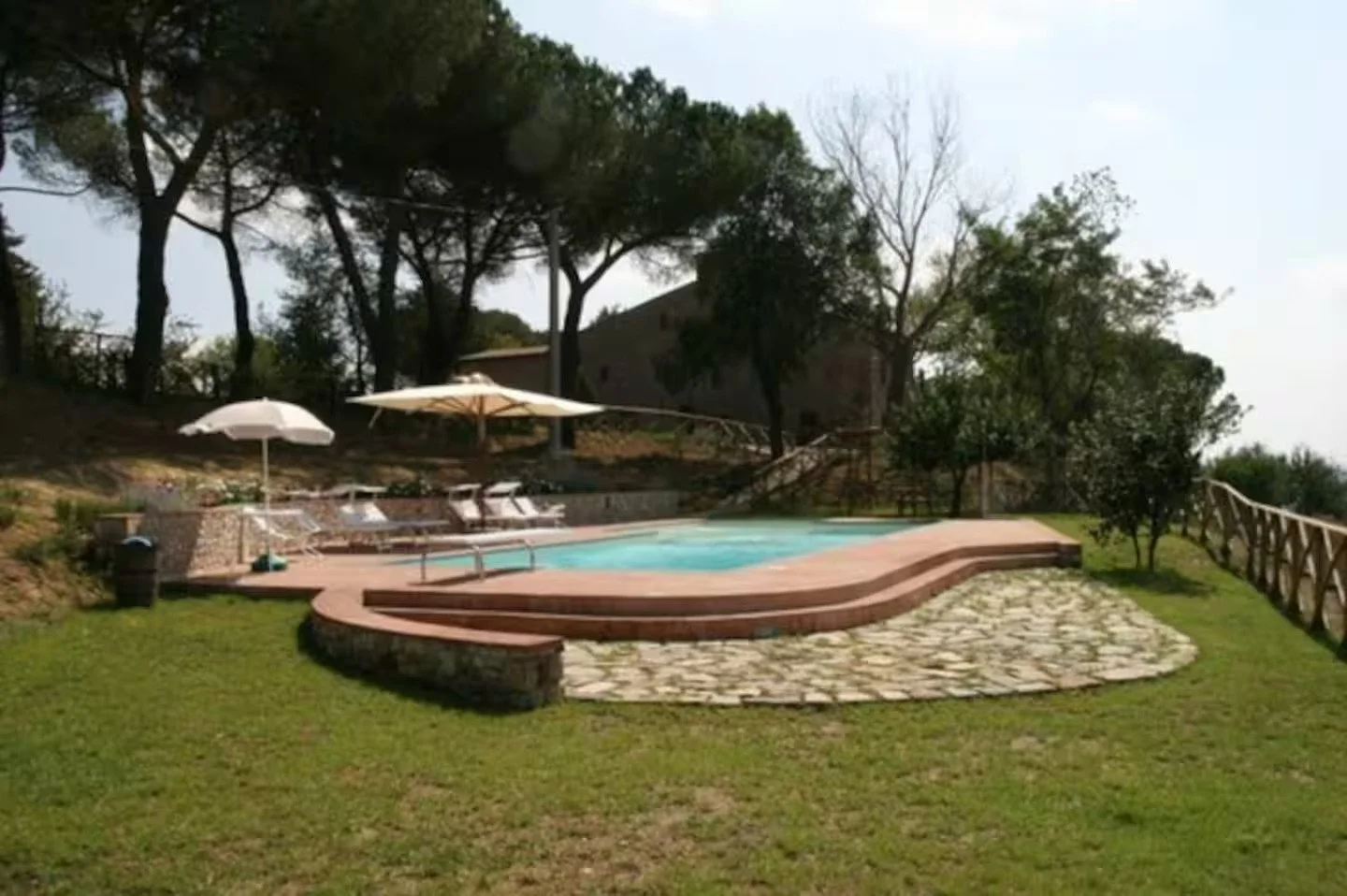 An outdoor swimming pool area with lounge chairs, umbrellas, and a surrounding grassy lawn, set against a backdrop of trees and a hillside.