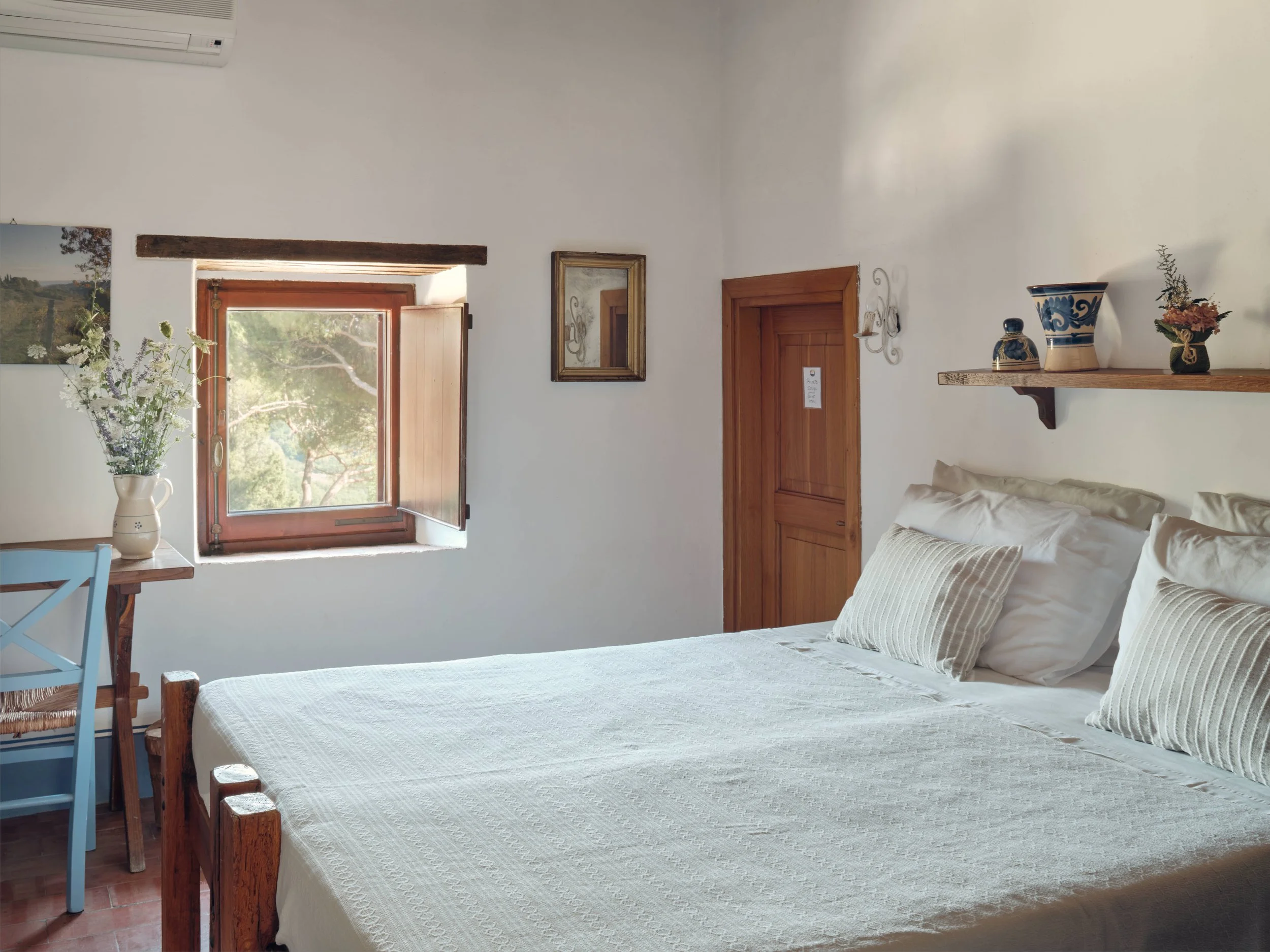 A cozy bedroom featuring a white bed with striped pillows, a wooden bedside table, a window with views of trees, and a simple wooden shelf with decorative vases and flower arrangements.