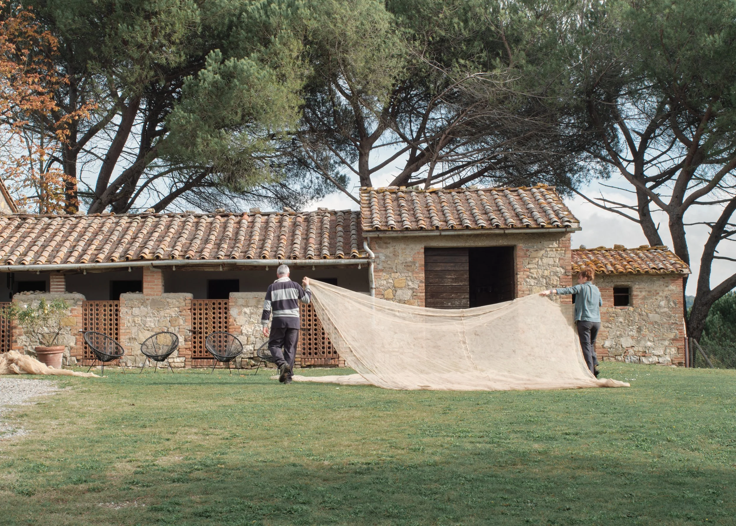 Two people setting up a large outdoor tent or cover on a grassy yard in front of a rustic brick and stone house with large trees in the background.