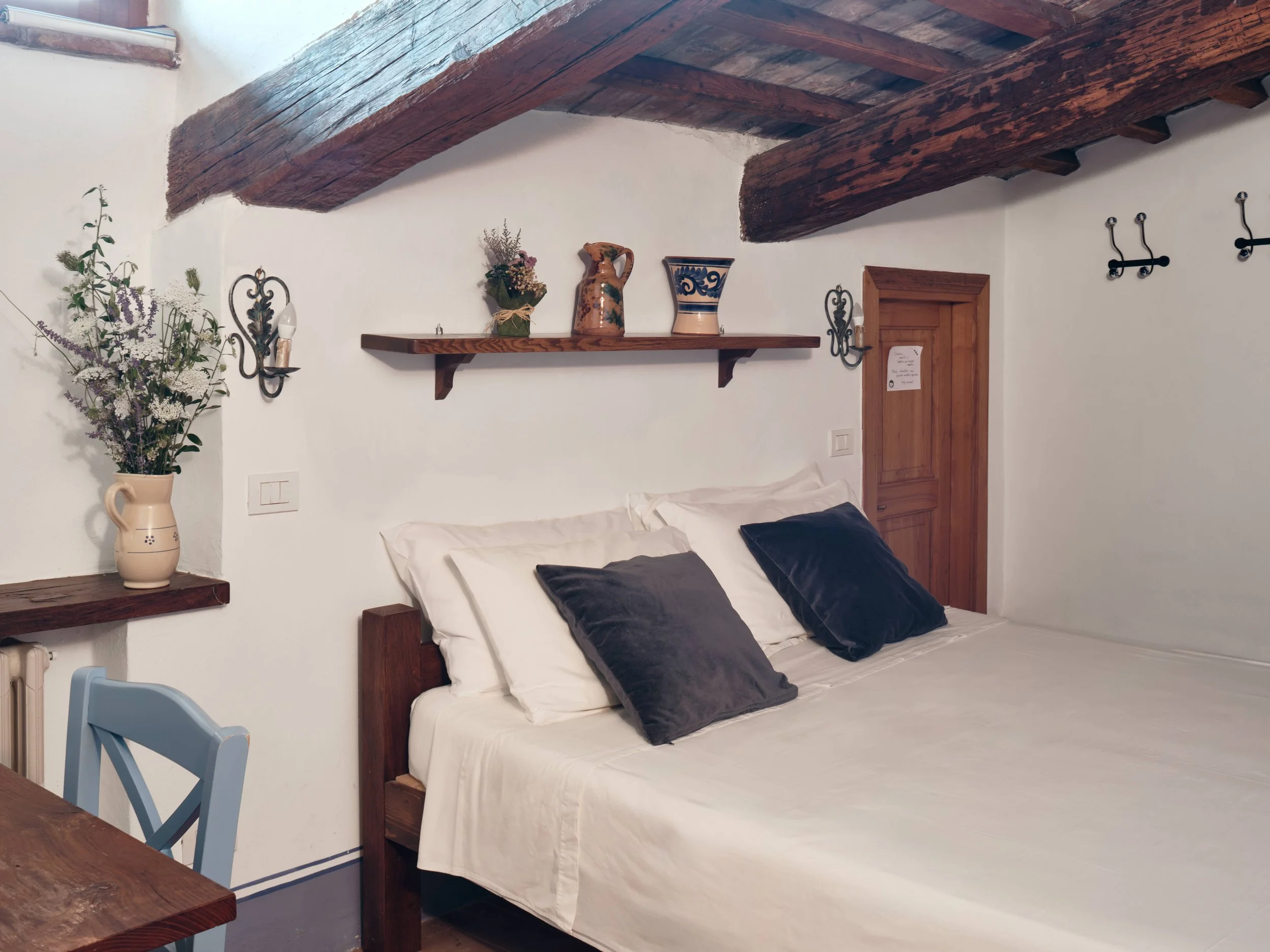 A cozy bedroom with a rustic wooden bed frame and white bedding with dark gray pillows. On the wall above, a simple wooden shelf holds decorative vases and a small bouquet of flowers. A vase with purple and white flowers is on a wooden nightstand to 