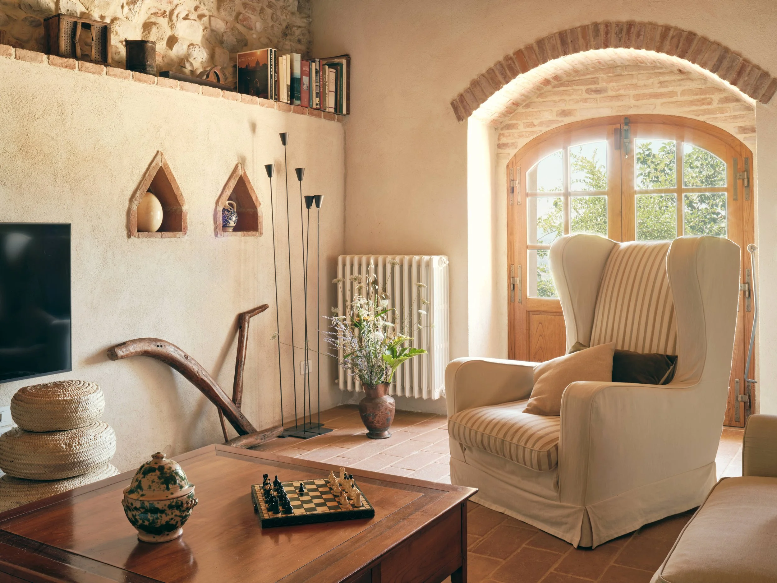 A cozy living room featuring a wooden coffee table with a decorative teapot and a chess set on it. Behind, there is a striped armchair, a tall white radiator, and large wooden-framed windows with views of greenery outside. The wall has built-in shelv