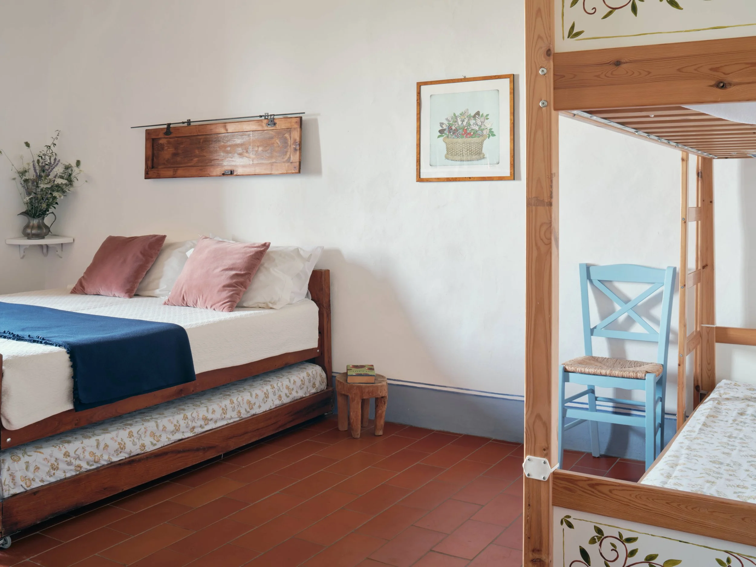 A bedroom with a wooden bed frame, pink pillows, a small side table with a book, a flower vase with flowers, and a picture of a basket of flowers on the wall. There is a wooden bunk bed frame and a blue chair.