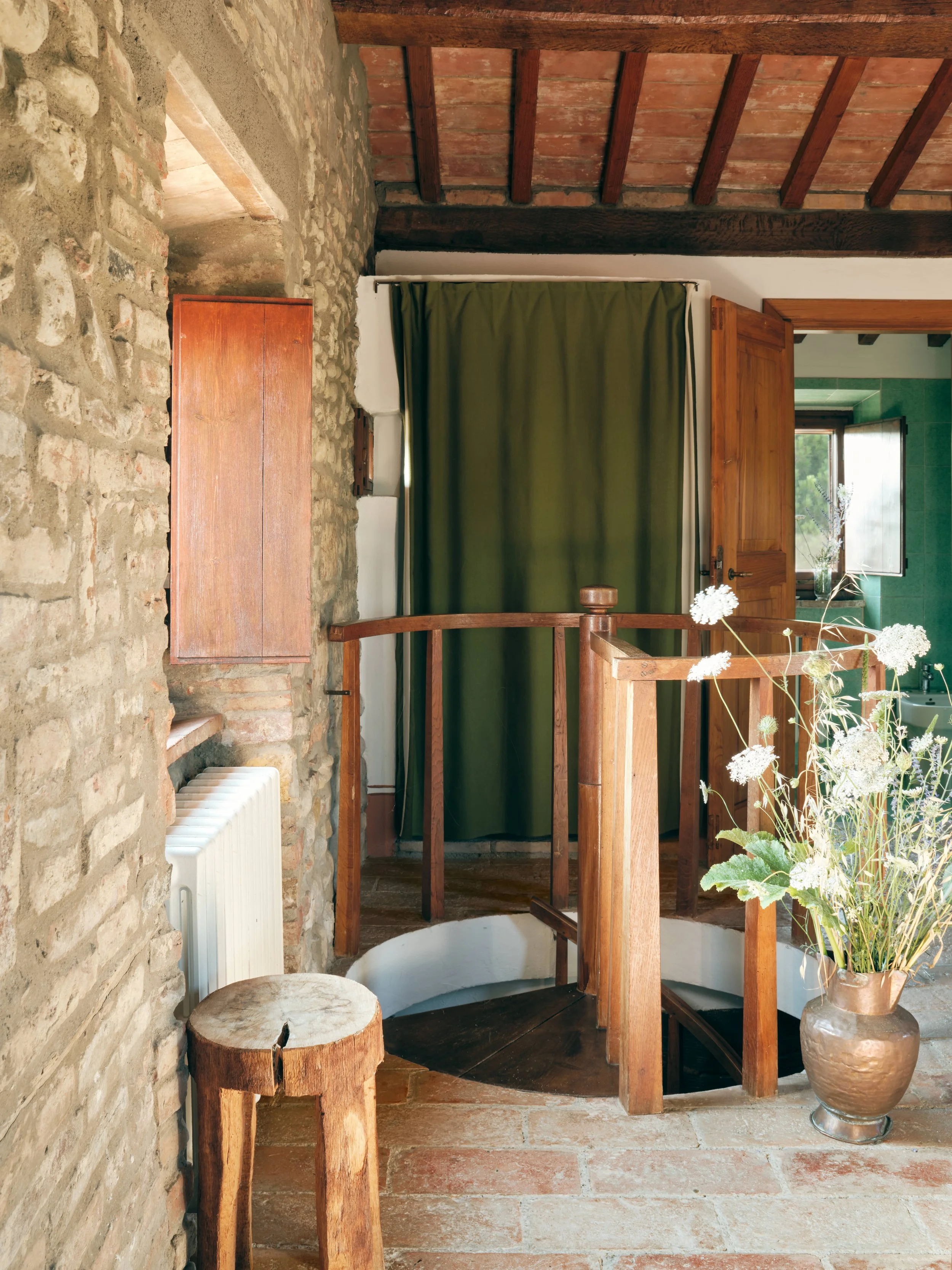 Interior view of a rustic room with a stone wall, wooden ceiling beams, green curtains, a wooden door, and a large vase with white flowers.