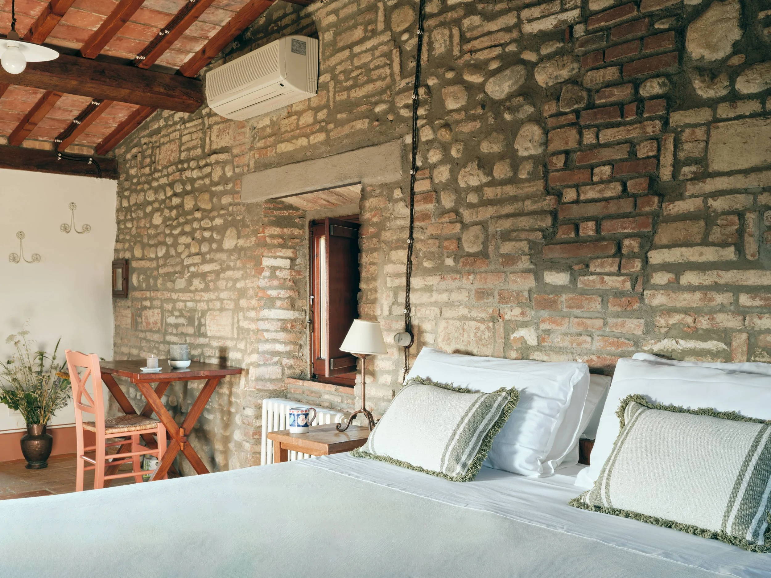A cozy living room with an exposed brick wall and wooden ceiling beams. There are white pillows on a sofa, a small wooden side table with a lamp and a mug, and a pink chair next to a small table with decorative items. A potted plant is in the corner,