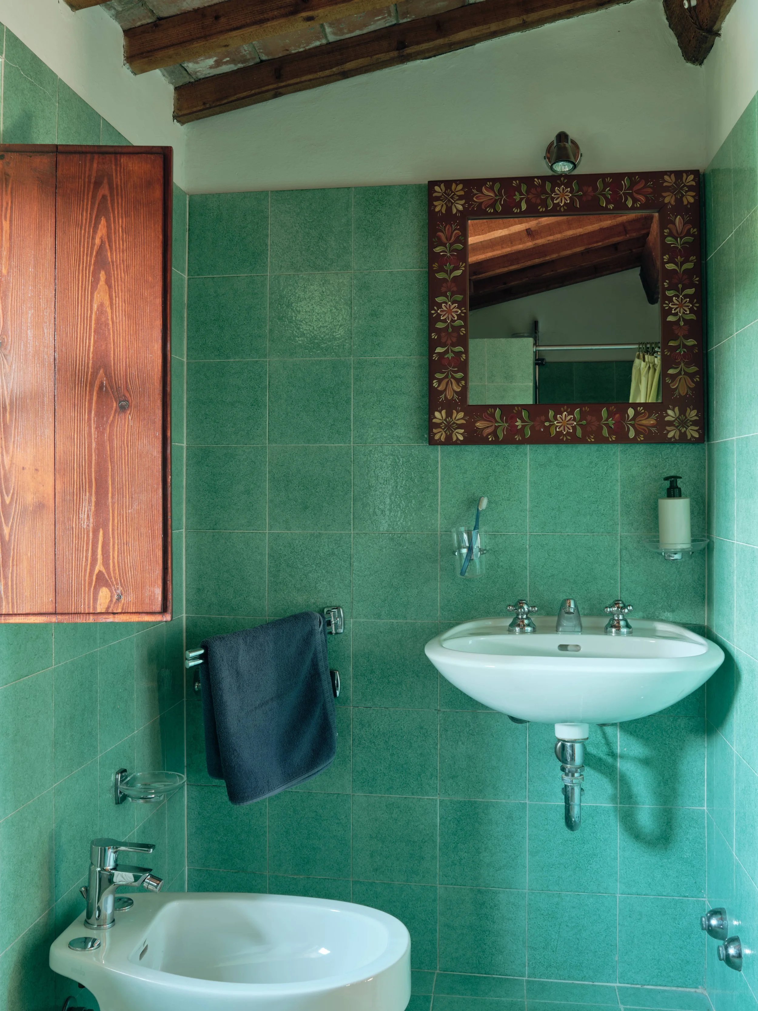 A green-tiled bathroom with a white sink and a mirror with a detailed floral frame. A small soap dispenser is next to the sink. A dark towel hangs on a rod, and a toothbrush is in a glass on a ledge. The ceiling has exposed wooden beams, and there is