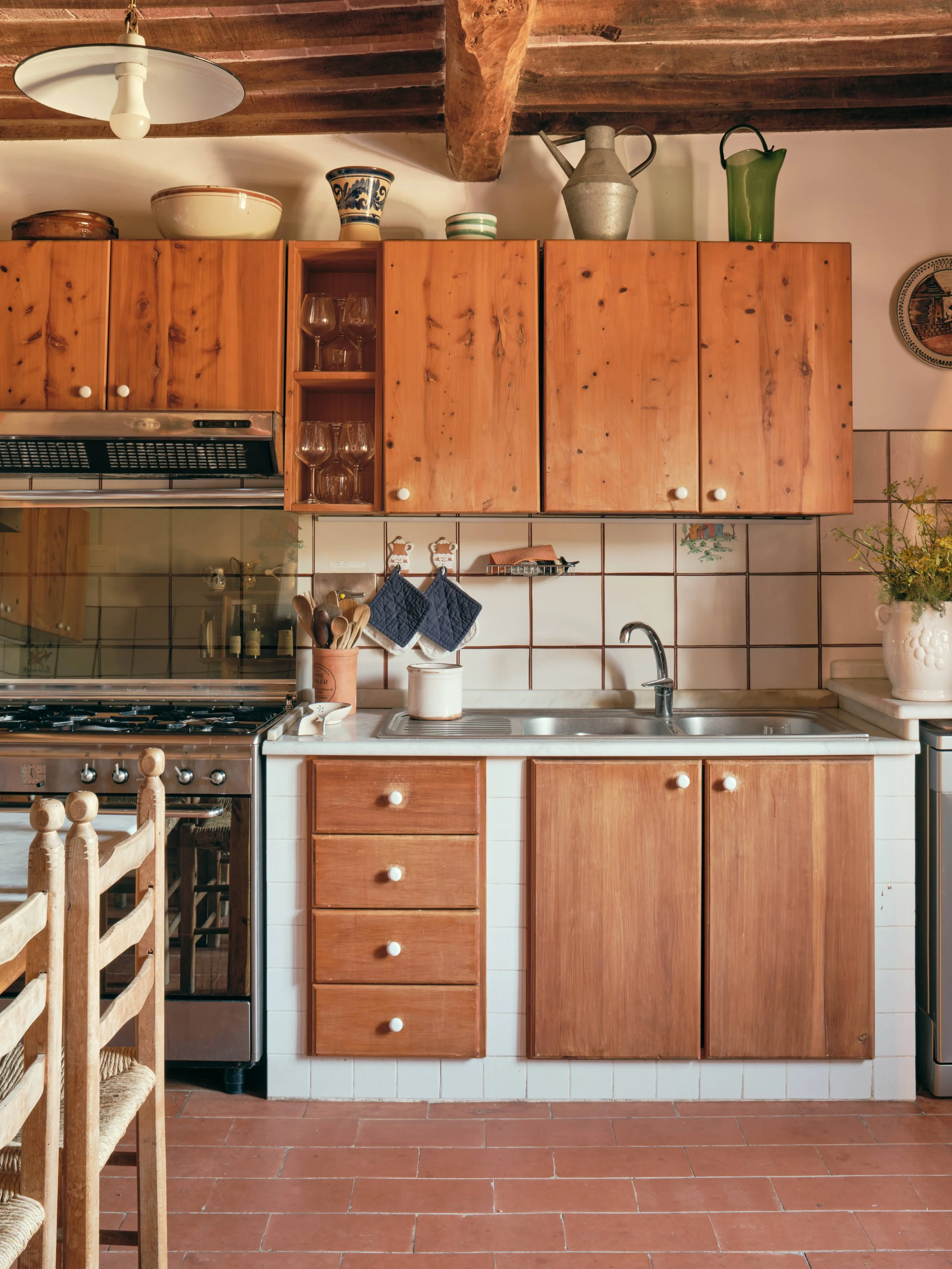 A kitchen with wooden cabinets, a white tiled backsplash, a gas stove, a sink, and decorative items on the upper cabinets including pottery, glassware, and a metal pitcher.