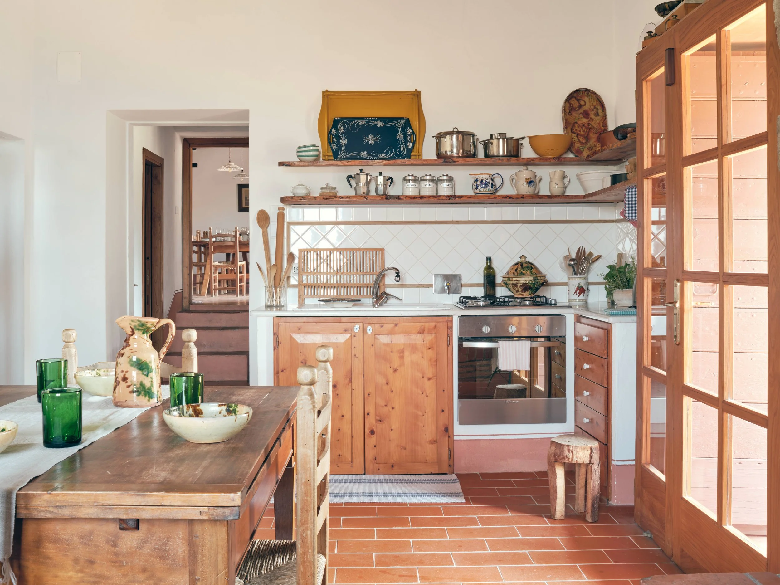 A cozy kitchen with wooden cabinets, an open wooden shelf with pots and dishes, a stovetop oven, and a dining table with green glasses and ceramic bowls.