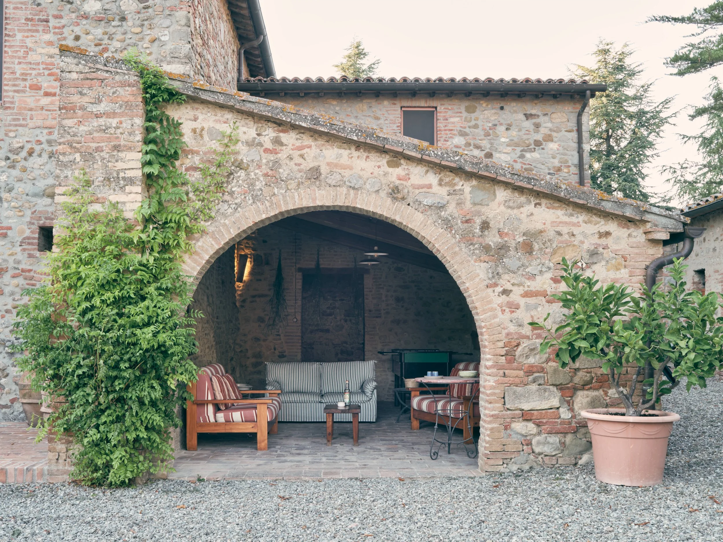 An outdoor patio area under an archway of a stone and brick building, with chairs and a table, potted plants, and trees in the background.