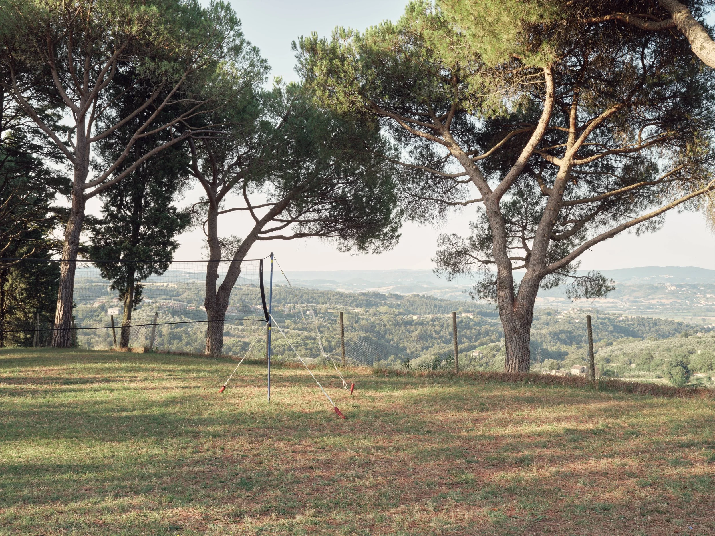 Tall pine trees overlooking a fenced grass yard with sports equipment, hills, and a town in the distance under a clear sky.