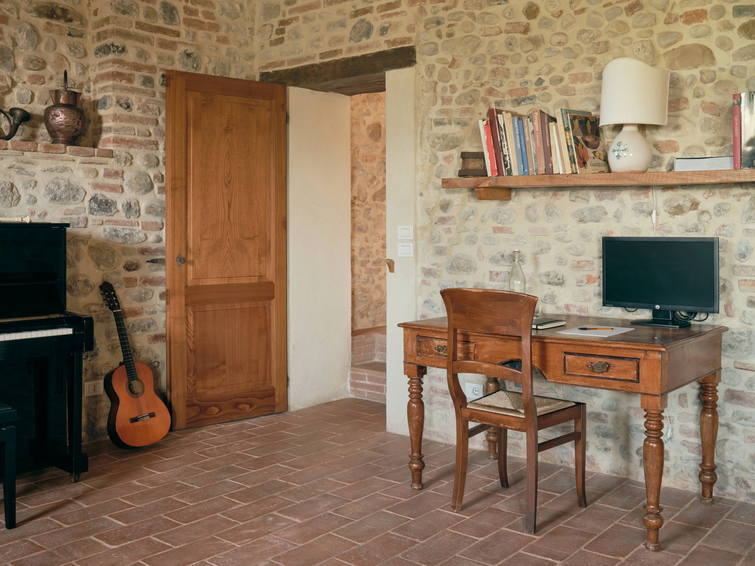 Interior room with stone walls, wooden door, brick floor, wooden desk with a chair, a small TV, bookshelf with books, a white lamp, and an acoustic guitar leaning against the wall.