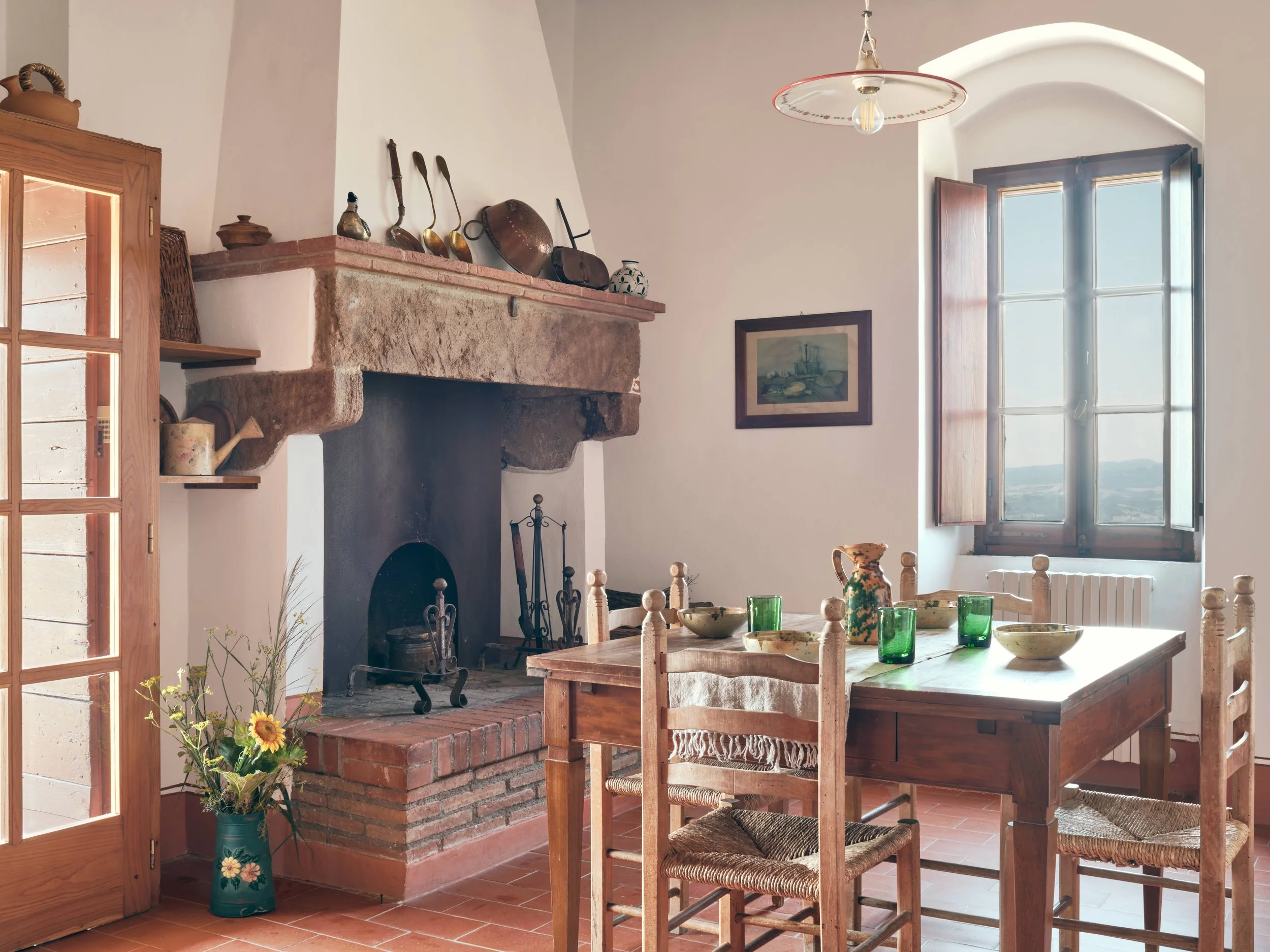 A rustic kitchen with a stone and brick fireplace, wooden dining table with six chairs, green glassware, bowls, a sunflowers in a vase, and a window with wooden shutters letting in natural light.